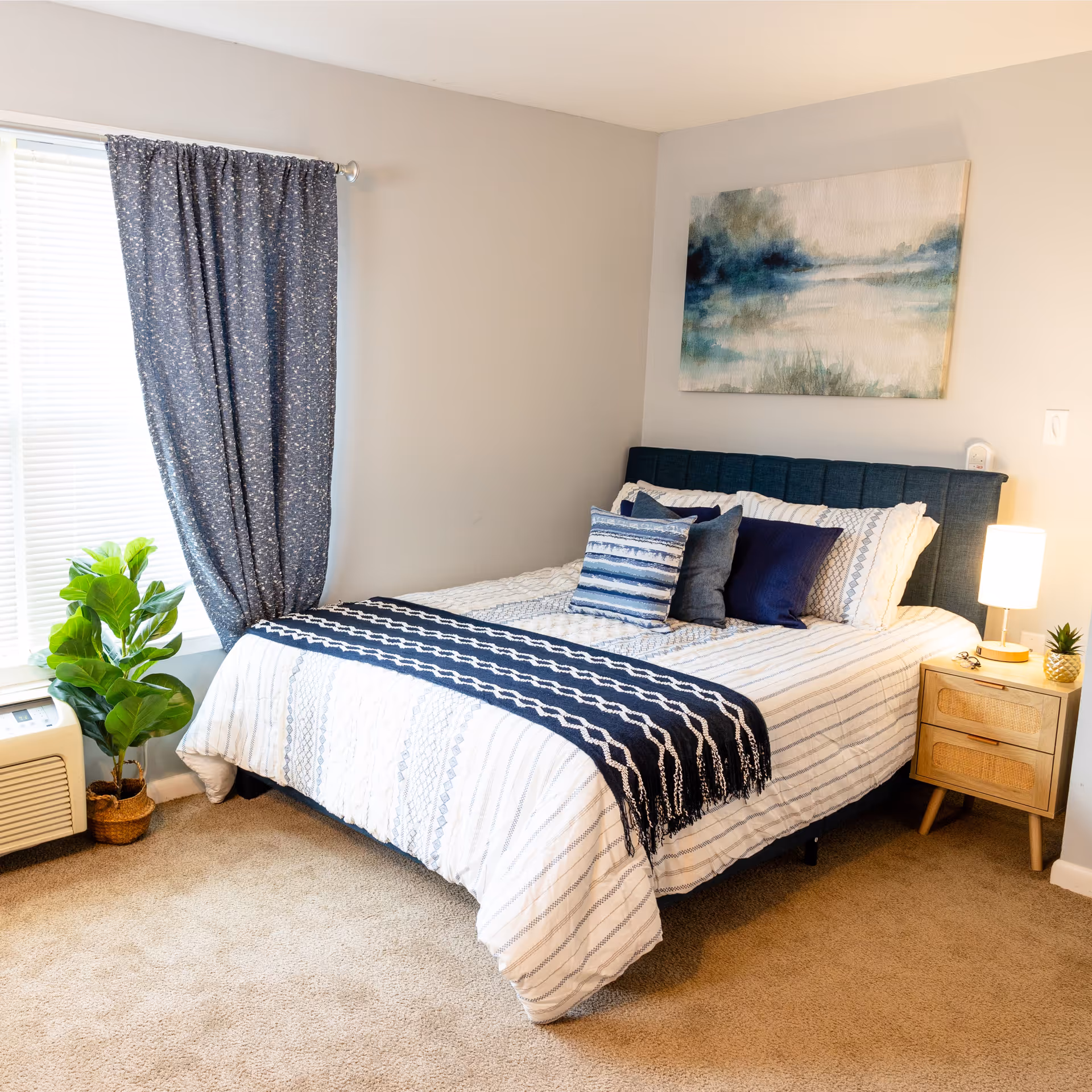 A neatly made bed with white and blue striped bedding and multiple decorative pillows in a bedroom. There is a wooden nightstand with a lamp and a small plant on it. A large window with blue curtains and a potted plant next to an air conditioning unit are visible. A landscape painting hangs on the wall above the bed.