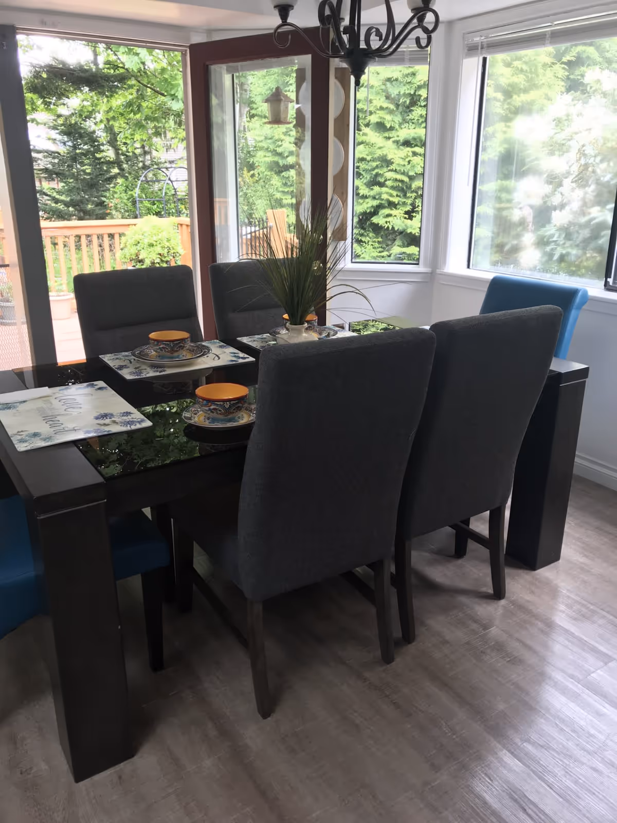 A dining area with a black glass-top table set for four with grey upholstered chairs and one blue chair. The table has placemats, plates, bowls, and a small plant centerpiece. Large windows and a glass door provide a view of green trees and a wooden deck outside.