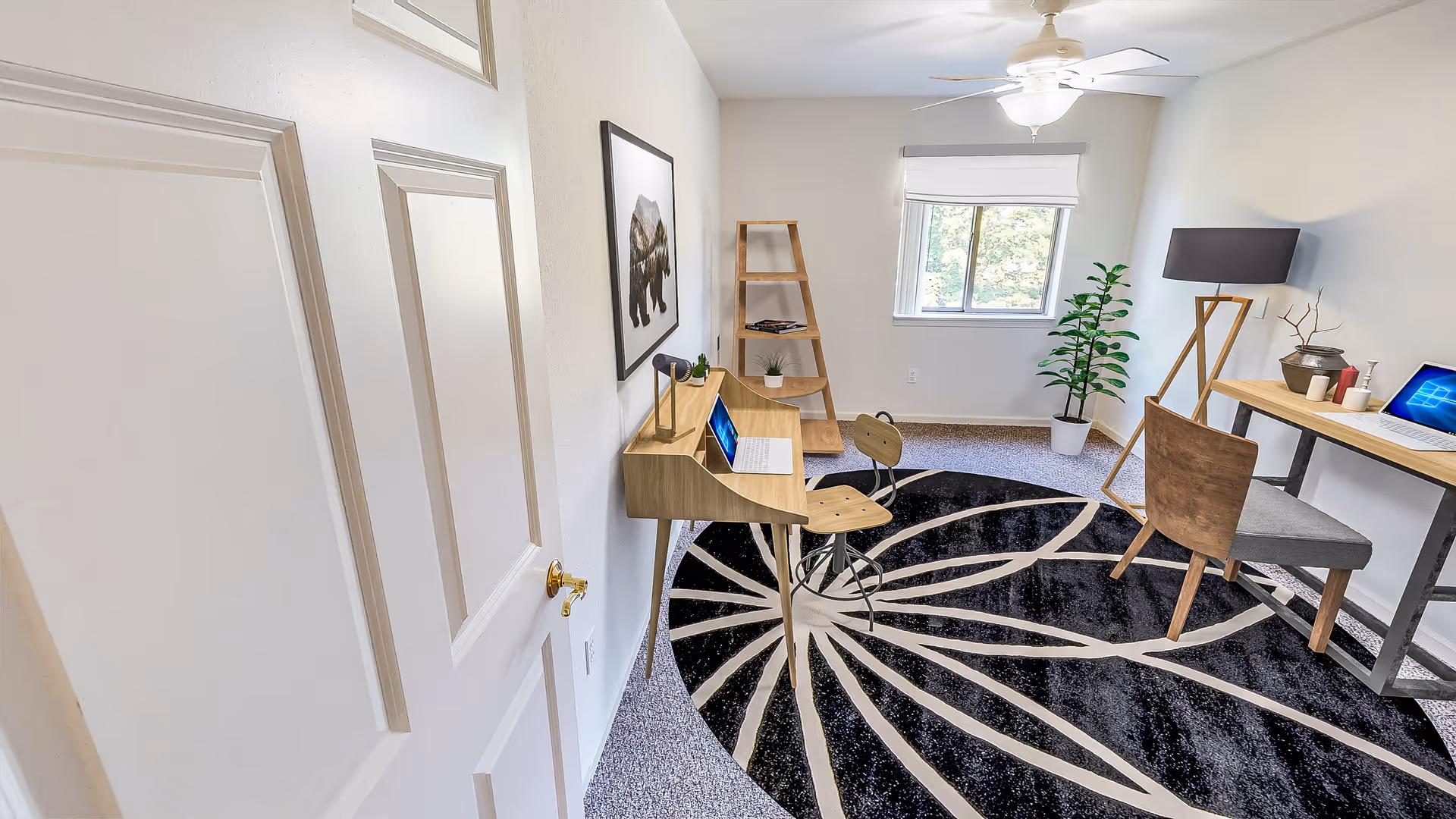 A bright room with two wooden desks, each with a laptop on top. There are two wooden chairs, a black and white patterned rug on the floor, a tall wooden shelf with plants and books, a potted plant near the window, a floor lamp, and a ceiling fan with a light. The walls are white, and there is a framed picture of a bear on the wall.