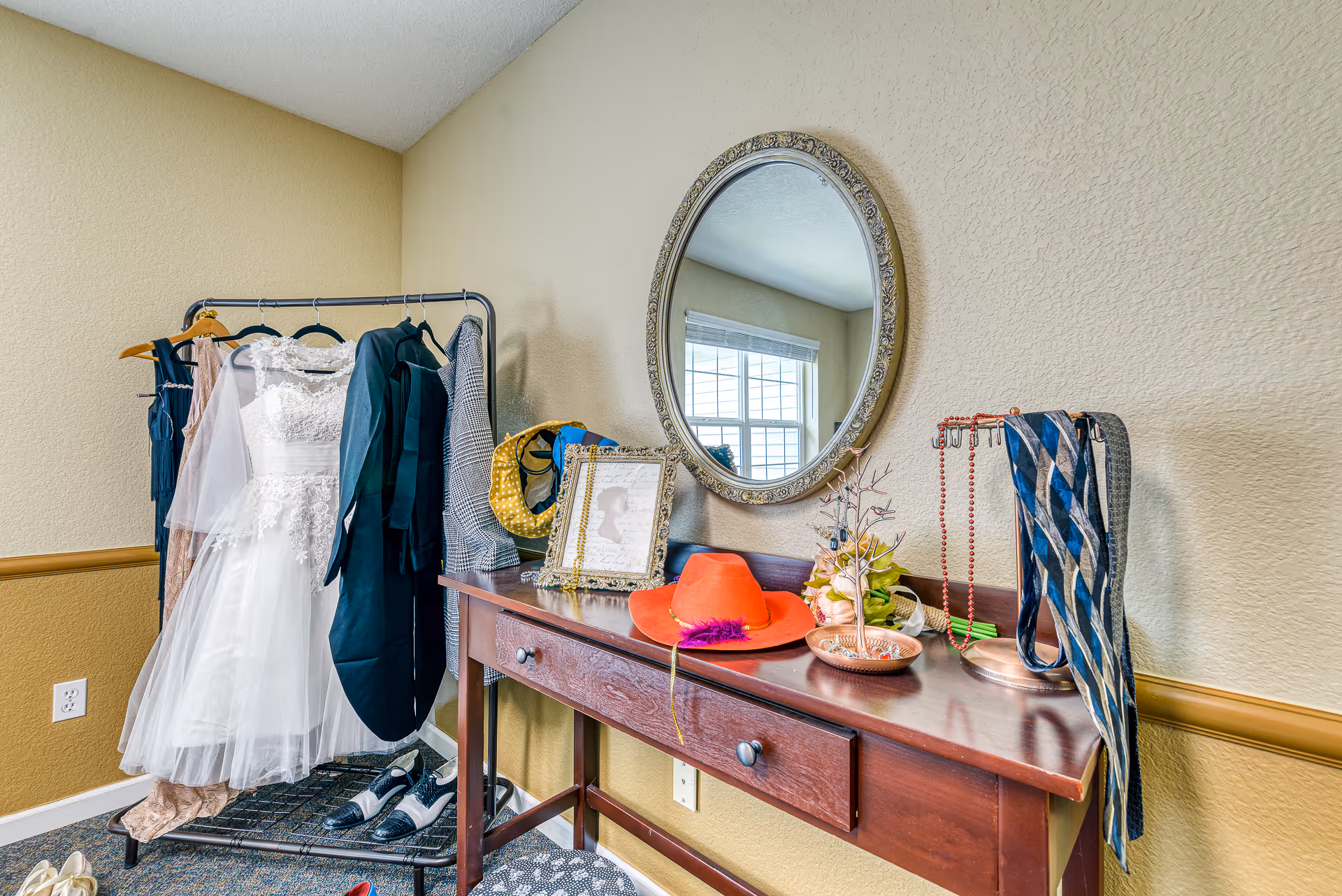 A dressing area with a clothing rack holding various garments including a white lace dress and a dark suit jacket. Below the rack are several pairs of shoes. Next to the rack is a wooden table with two drawers, topped with a round ornate mirror, a red hat with a purple feather, a framed silhouette picture, a jewelry tree with necklaces, and a striped scarf hanging on a stand. The walls are painted beige with a textured finish.