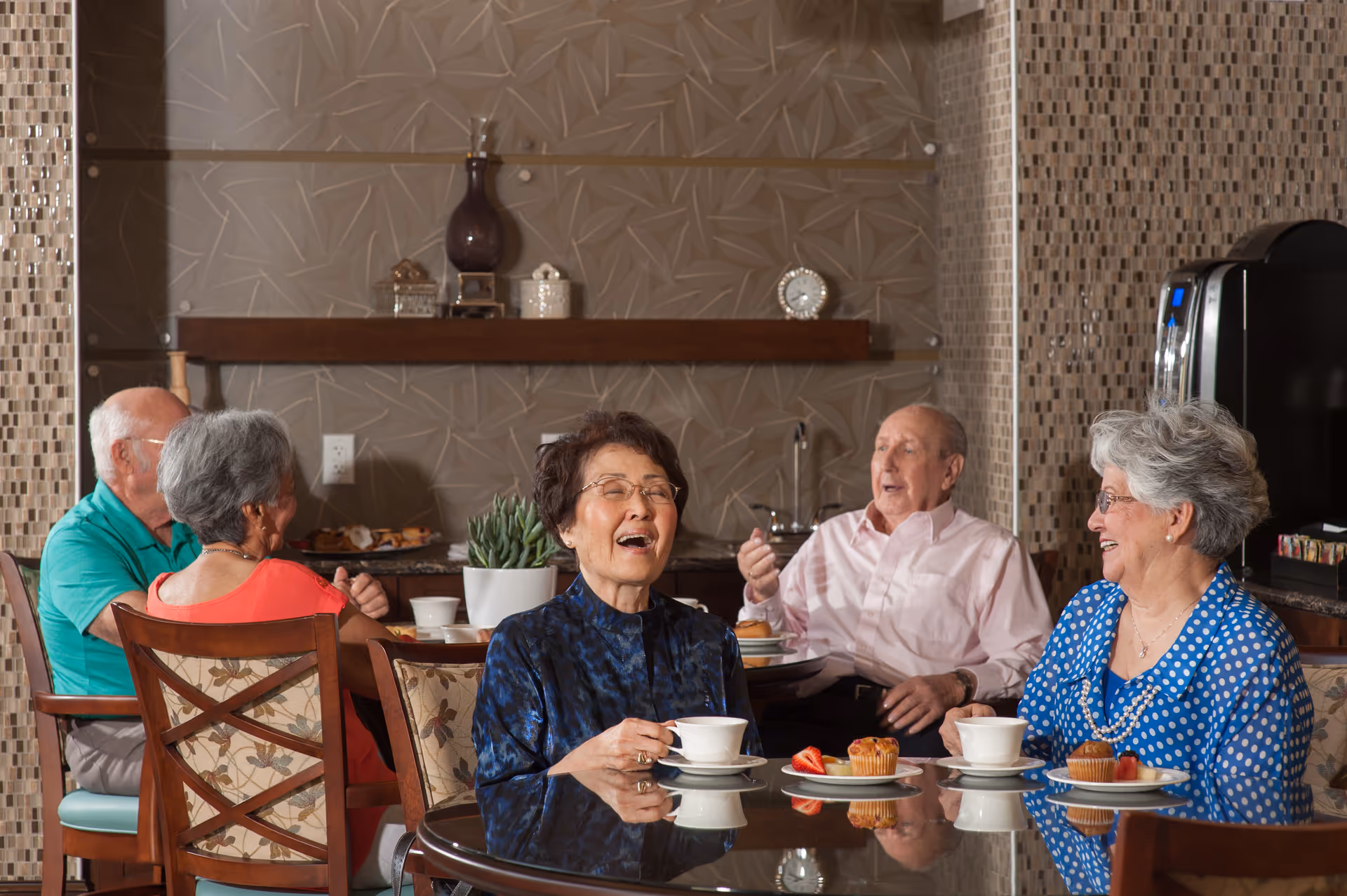 A group of elderly residents sit around a table in a communal dining area, enjoying coffee, pastries, and conversation.