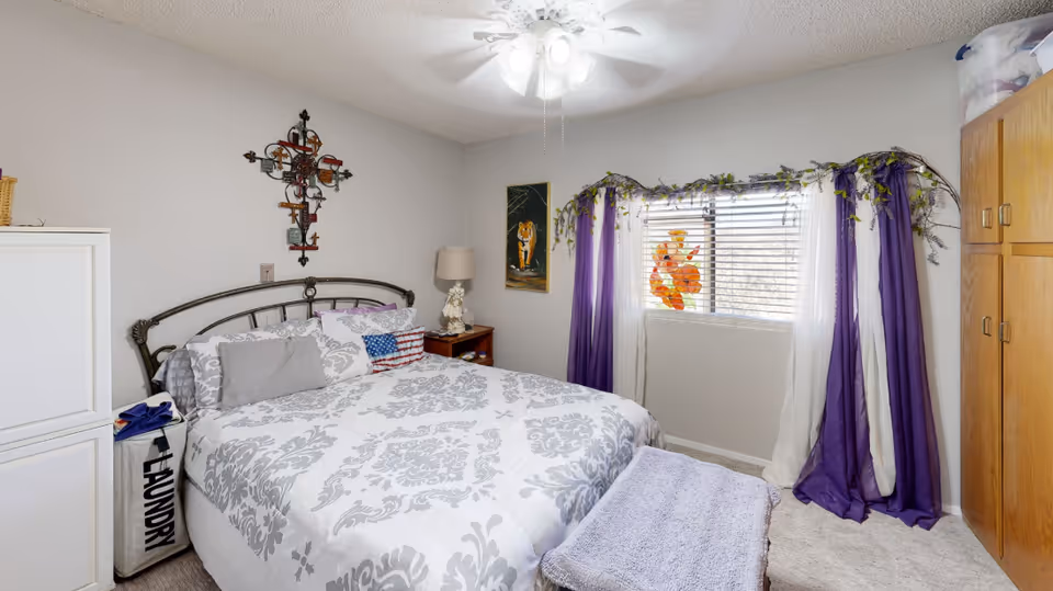 A neatly decorated bedroom with a bed, nightstand, purple curtains over a window, and a ceiling fan.