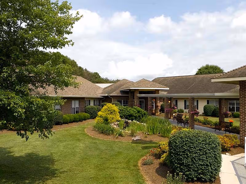 Exterior view of Knoxville Landing - A Willow Ridge Senior Living community showing a single-story brick building surrounded by well-maintained landscaping including bushes, trees, and flower beds under a partly cloudy sky.