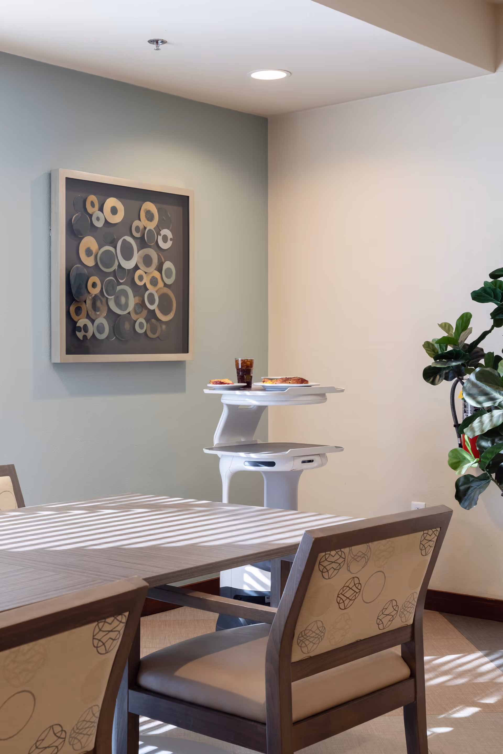 A dining area with a wooden table and cushioned chairs featuring a circular pattern. On the wall is a framed abstract artwork with various circular shapes. A white serving cart holds plates with food and a glass of beverage. A green leafy plant is visible on the right side.