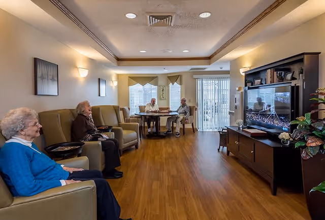 A cozy senior living common area with three elderly women seated on armchairs on the left side and two elderly men sitting at a round table near large windows with blinds. A large flat-screen TV is on the right side atop a dark wooden cabinet, and the room has wooden flooring and soft lighting.