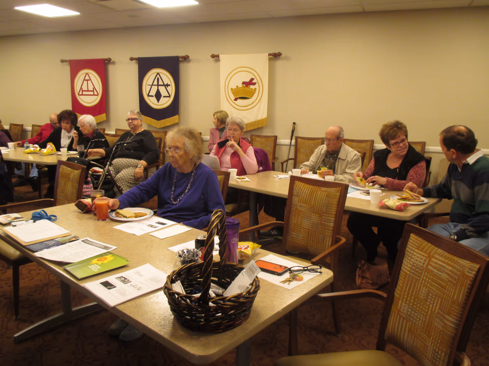 A group of elderly people sitting around tables in a meeting or dining room, eating and conversing. The room has beige walls with three banners hanging on the wall behind them, each with different symbols. There are papers, a basket, and some personal items on the tables.
