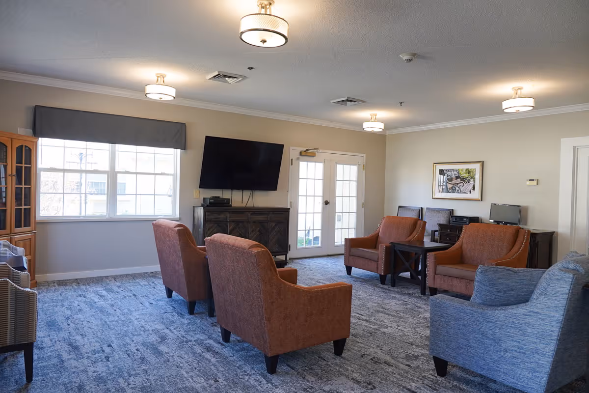 Bright communal lounge with multiple armchairs, a wall-mounted TV, side tables, and French doors.