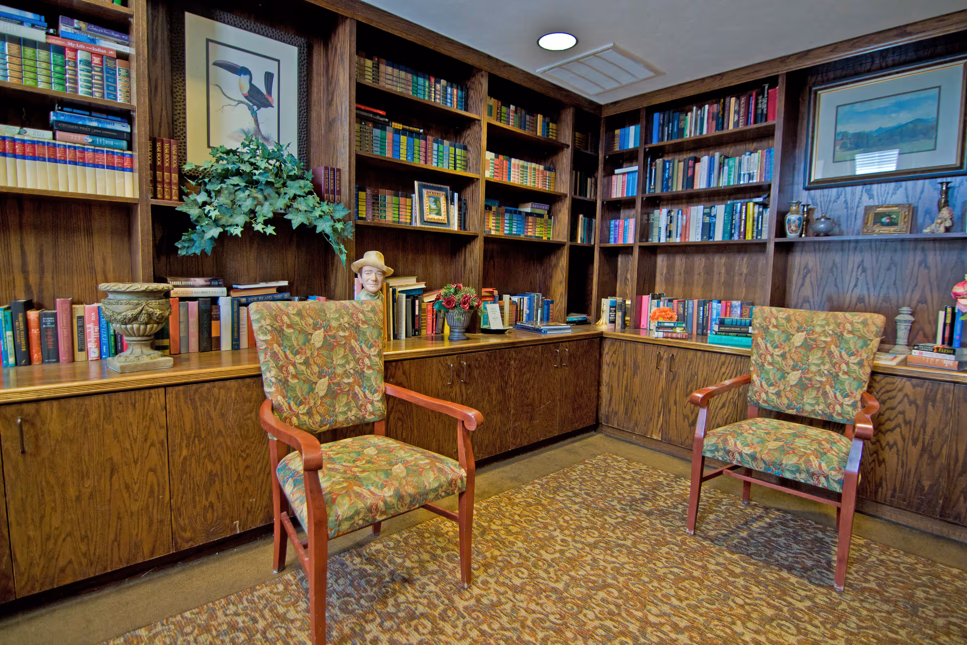 A cozy library room with wooden bookshelves filled with books and decorative items. Two floral upholstered armchairs with wooden arms are placed on a patterned carpet in front of the shelves. The room has warm lighting and framed artwork on the walls.