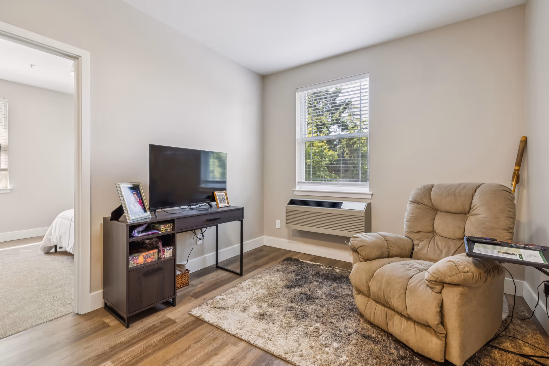 A cozy living room with a beige recliner chair next to a small side table holding a remote and a book. Across from the chair is a dark wooden TV stand with a flat-screen television and framed photos. A window with white blinds lets in natural light, and a beige and brown area rug covers part of the wooden floor. An open doorway leads to a bedroom with a bed visible.