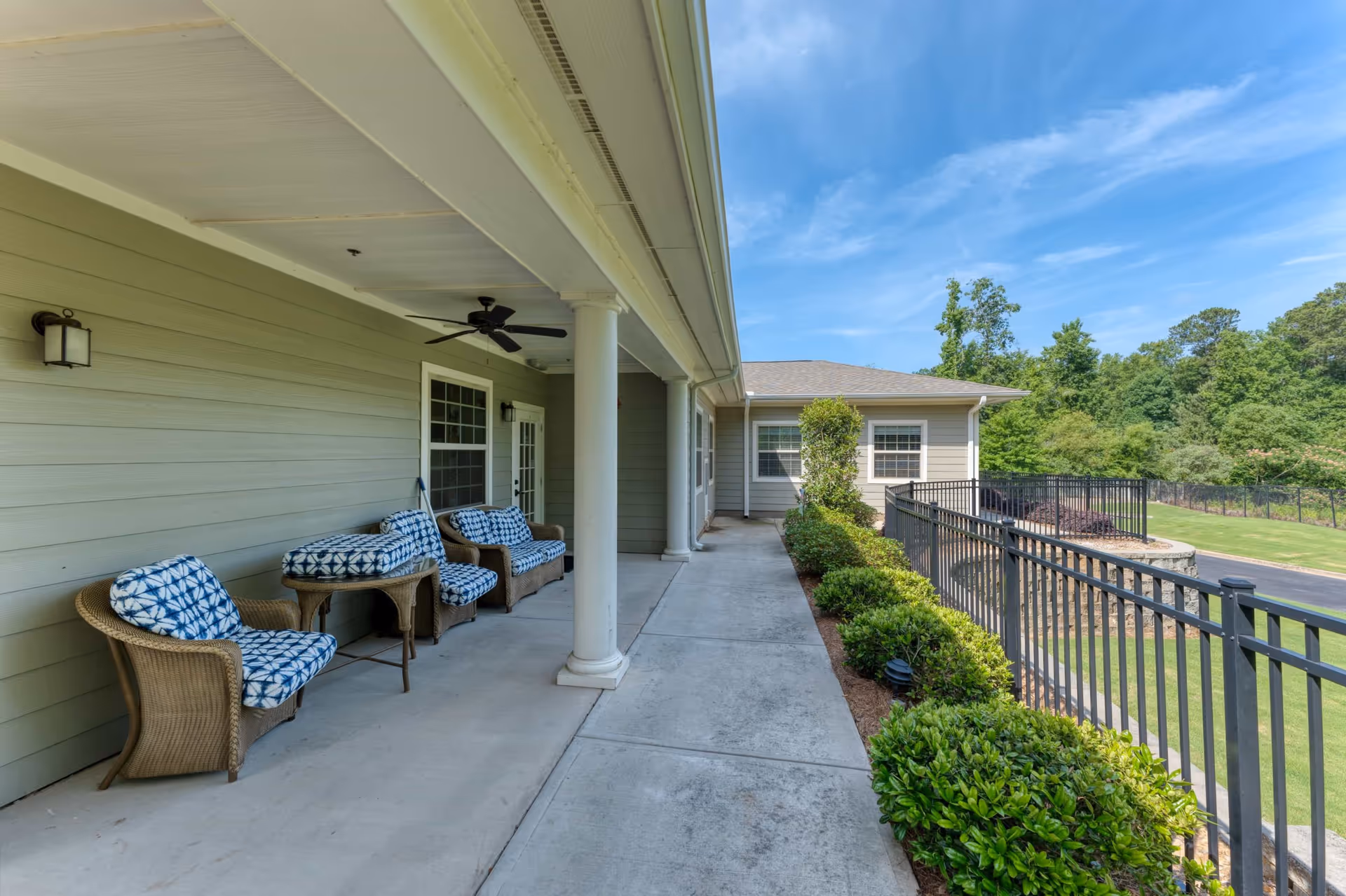 Covered outdoor patio area with wicker chairs and blue patterned cushions, a ceiling fan, and a concrete walkway next to a building with light green siding. There is a black metal fence and landscaped bushes along the walkway, with a view of trees and a blue sky in the background.