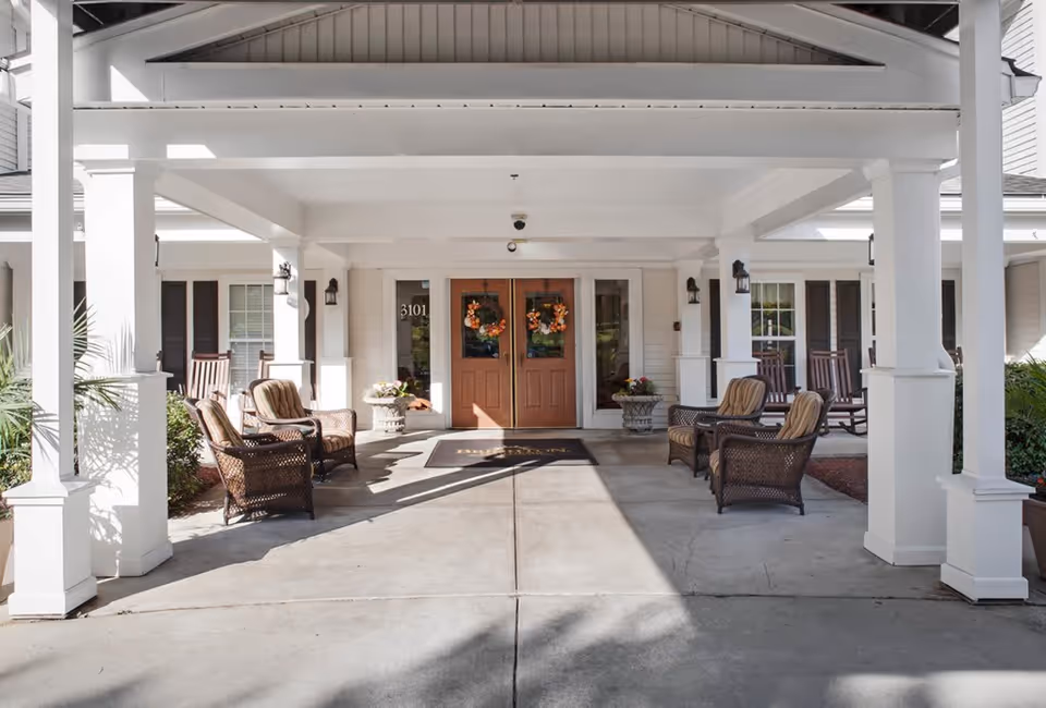 Covered entrance area of a senior living facility with white pillars and ceiling. There are four cushioned wicker chairs arranged in pairs on either side of the entrance. Two wooden double doors with autumn wreaths are centered in the background, flanked by windows and potted plants. The building exterior is white with black shutters and rocking chairs visible on the porch.