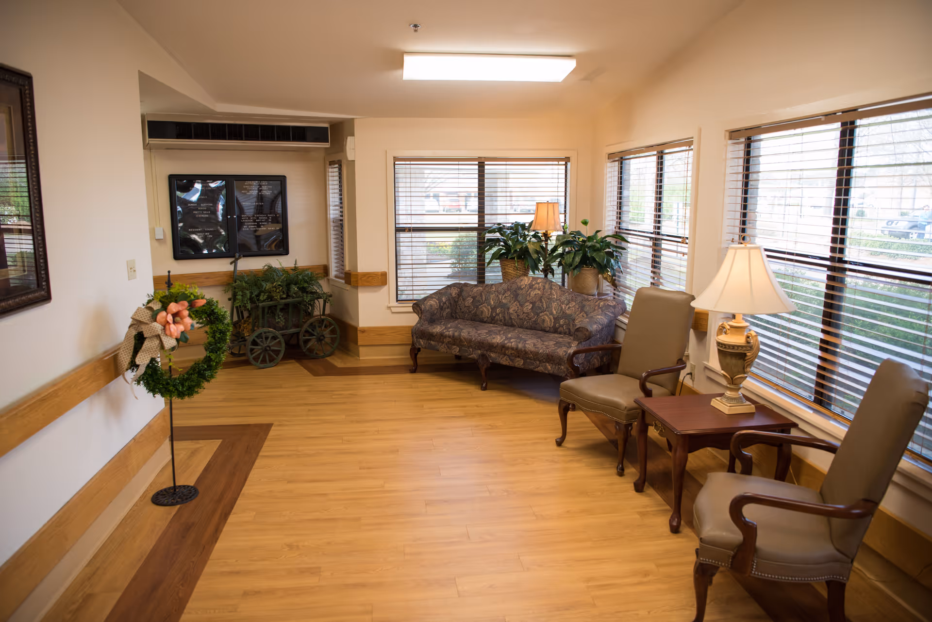 A cozy sitting area in The Oaks Center with a patterned sofa, two armchairs, a wooden side table with a lamp, several potted plants, and large windows with blinds letting in natural light. The floor is wood with a decorative inlay, and there is a decorative wreath on a stand near the wall.