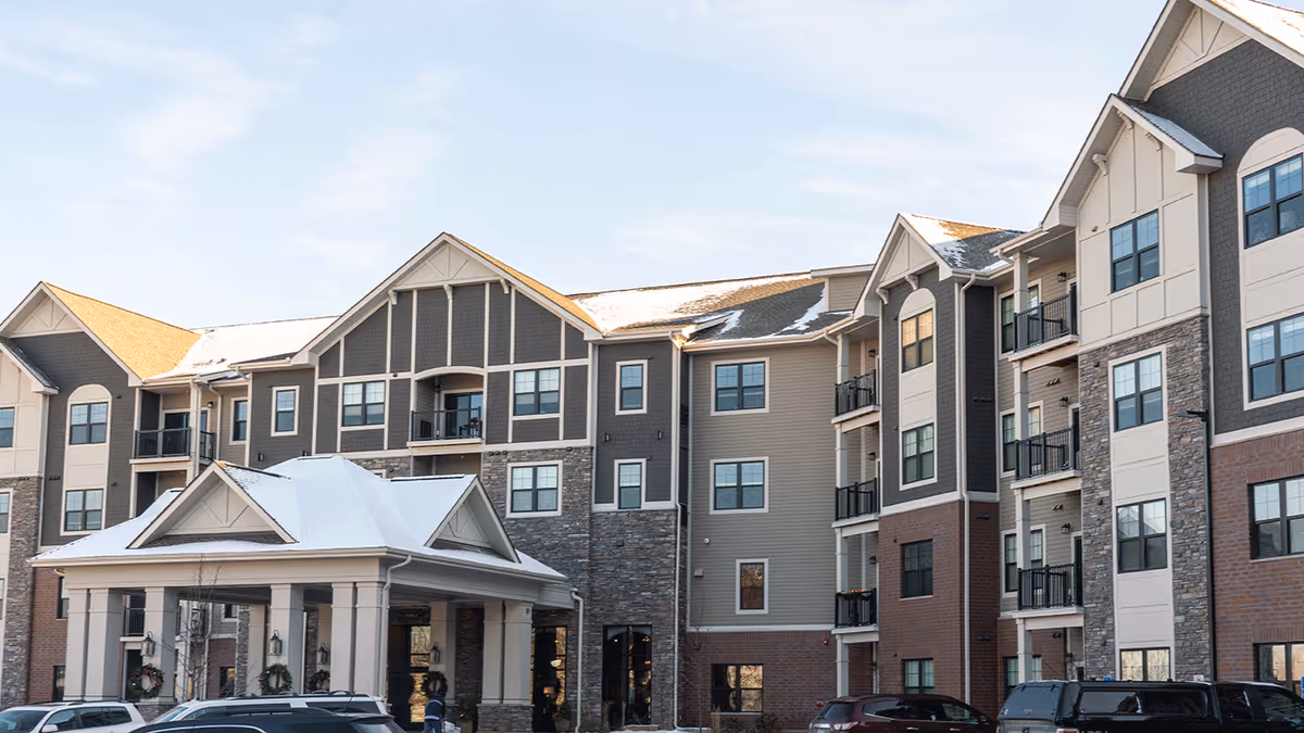 Exterior view of a multi-story senior living facility building with a covered entrance, multiple windows, balconies, and a mix of brick, stone, and siding materials under a partly cloudy sky.