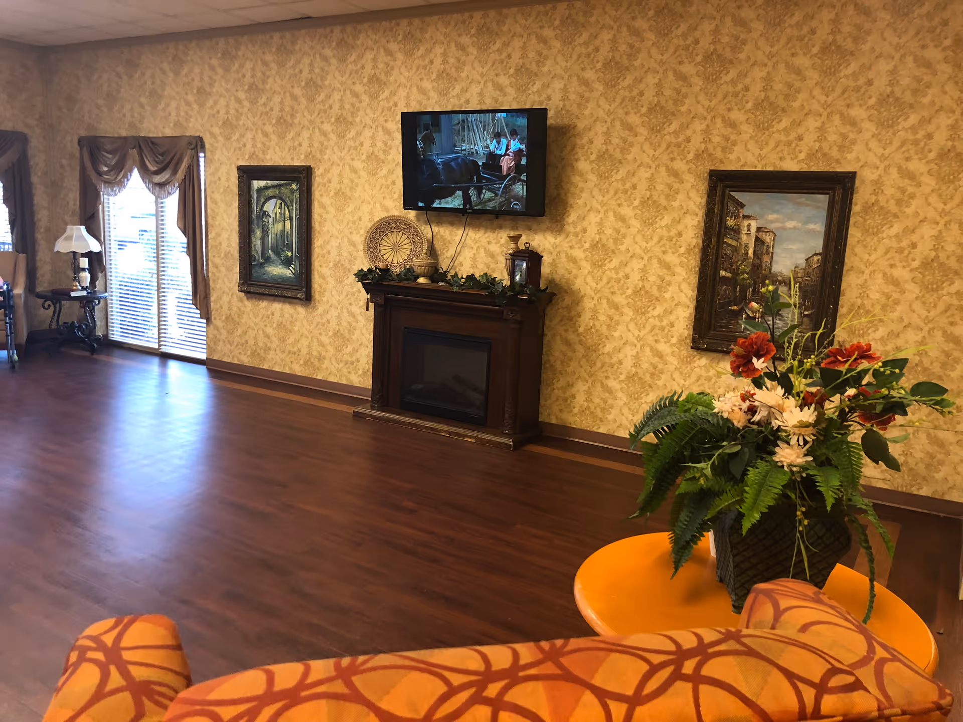 A cozy living room with wooden flooring, patterned wallpaper, and a mounted flat-screen TV above a dark wooden electric fireplace. The room features framed paintings on the walls, a side table with a lamp near a window with draped curtains, and a round orange table with a floral arrangement in the foreground.