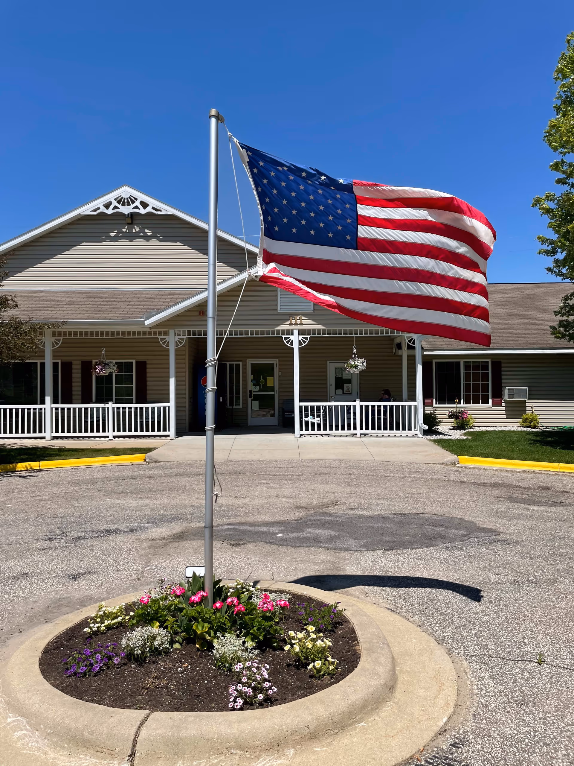 American flag flying over a circular flower bed in front of a building entrance with a covered porch.