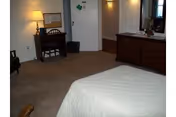 A bedroom with a bed covered in a white quilt, a wooden dresser with a mirror, a small desk with a lamp and chair, and a door with a shamrock decoration. The room has beige carpet and white walls.