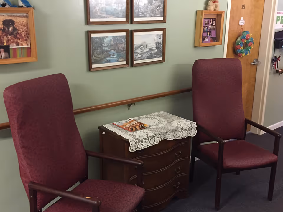 Two maroon upholstered chairs with wooden arms are placed on either side of a small wooden chest of drawers covered with a lace doily and a magazine. The wall behind is painted light green and decorated with framed pictures and a bulletin board. A wooden door with a colorful wreath and the number 15 is visible in the background.