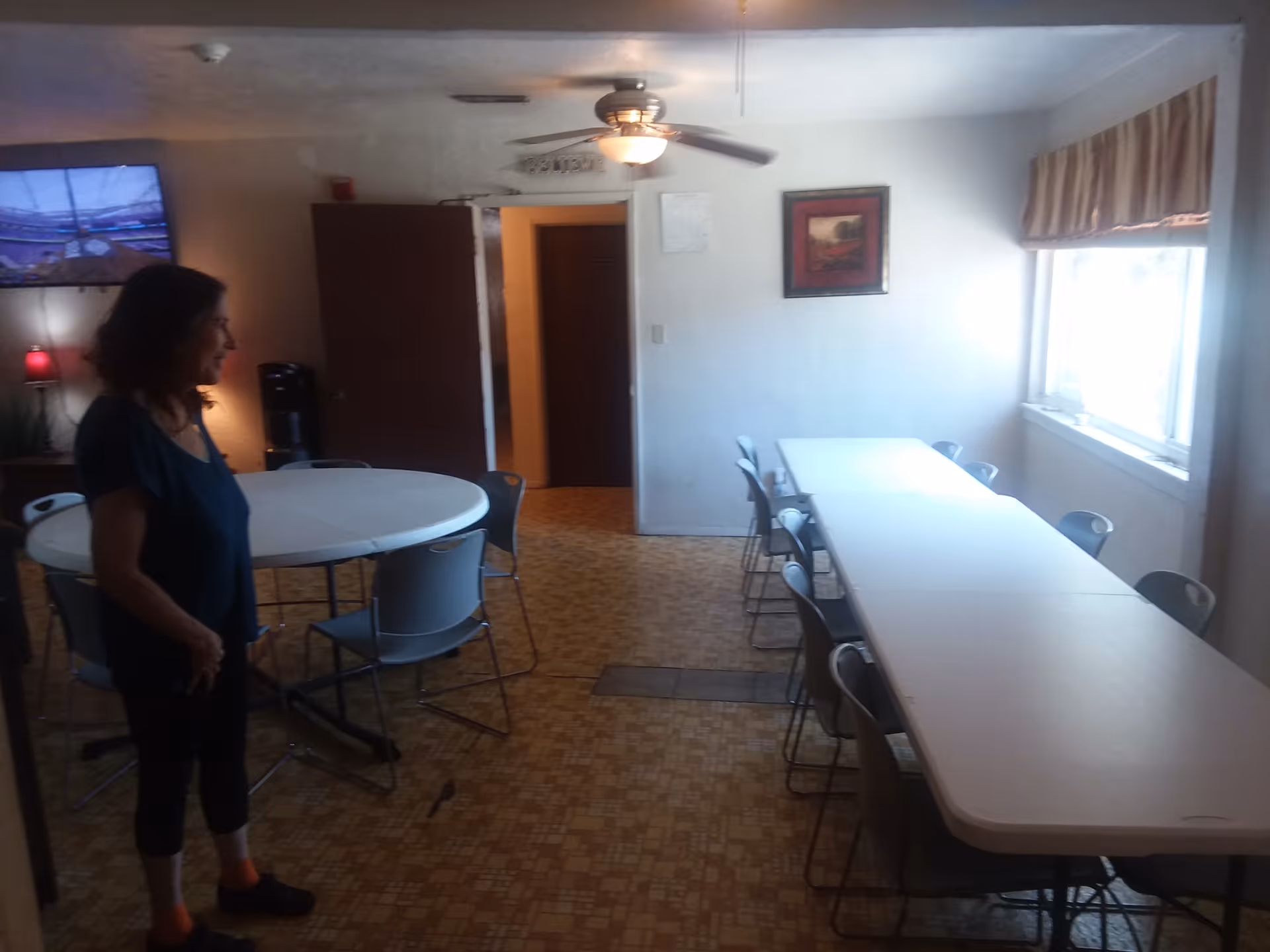 A dining room with two tables, one round and one rectangular, surrounded by chairs. A woman stands near the round table. There is a ceiling fan with a light, a window with curtains, a painting on the wall, and a television mounted on the wall showing a sports event.