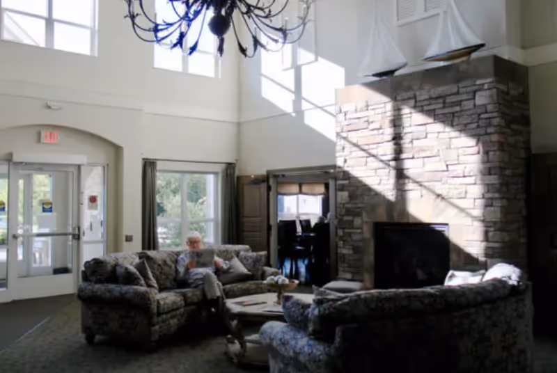 A bright and spacious senior living facility common area with high ceilings, large windows, and a stone fireplace. There are two patterned sofas arranged around a coffee table, and a person is sitting on one of the sofas reading. A chandelier hangs from the ceiling, and there is a doorway leading to another room with a dining table and chairs.