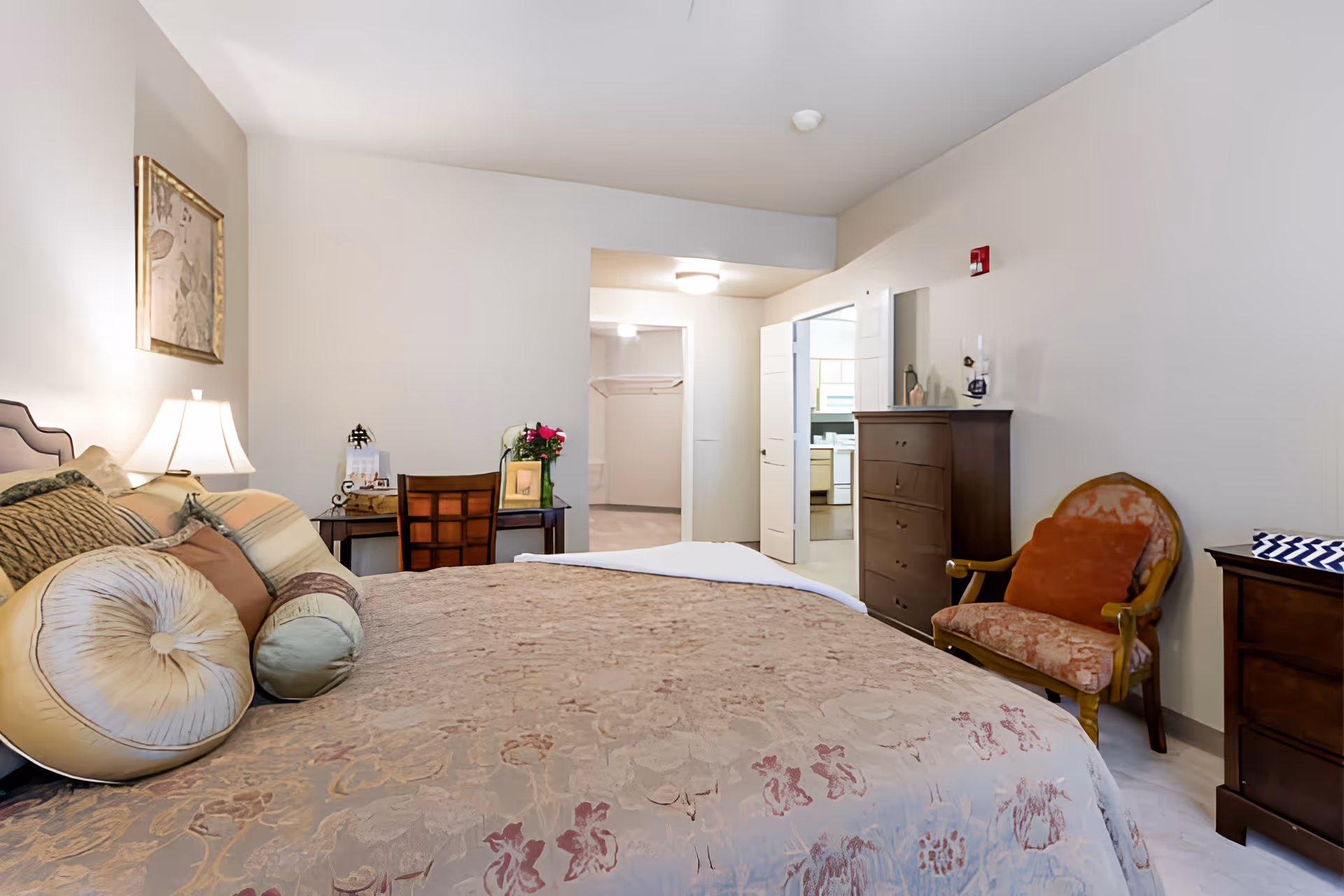 Spacious furnished bedroom with a large bed and decorative pillows, a bedside lamp, dresser and chairs, with an open doorway to an adjacent room.