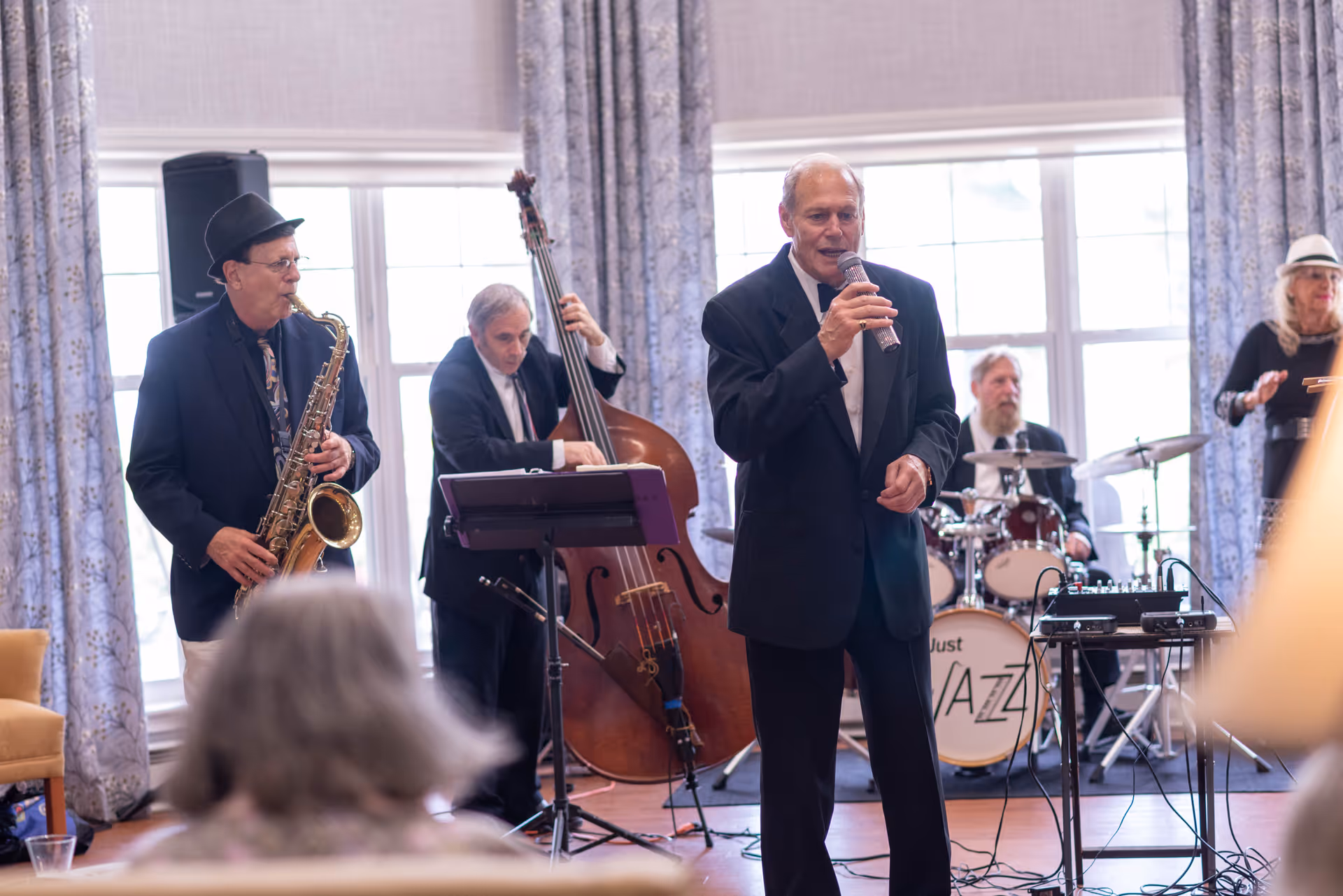 A jazz band performing indoors with a saxophonist, double bassist, drummer, and a singer holding a microphone. The room has large windows with patterned curtains and an audience member is visible in the foreground.