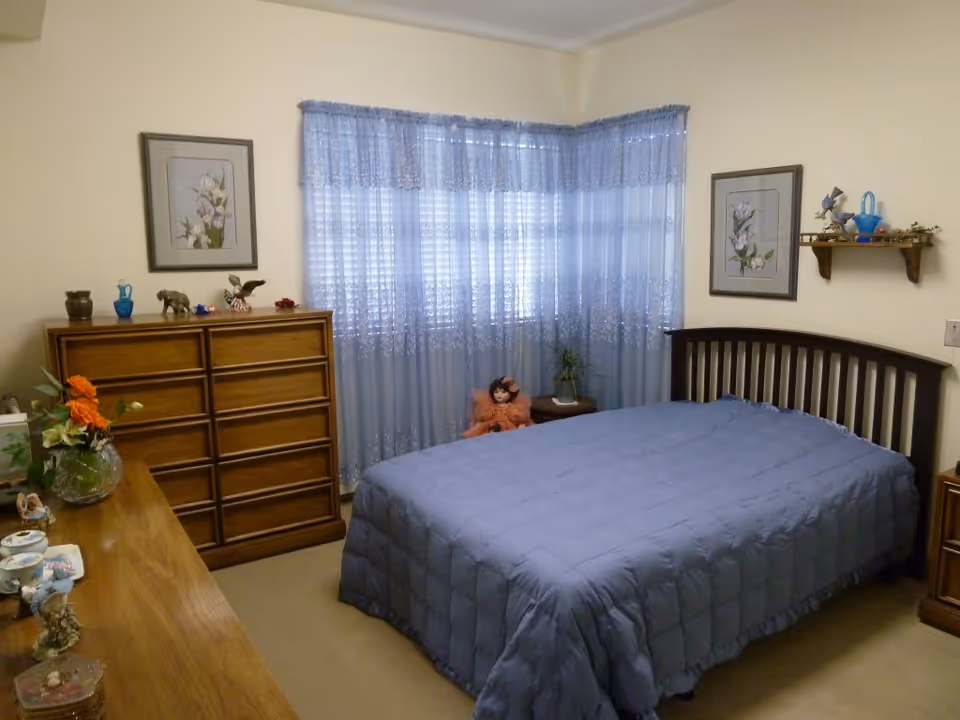 Tidy bedroom with a blue-covered bed, wooden dressers and nightstand, blue lace curtains, and wall-mounted artwork.