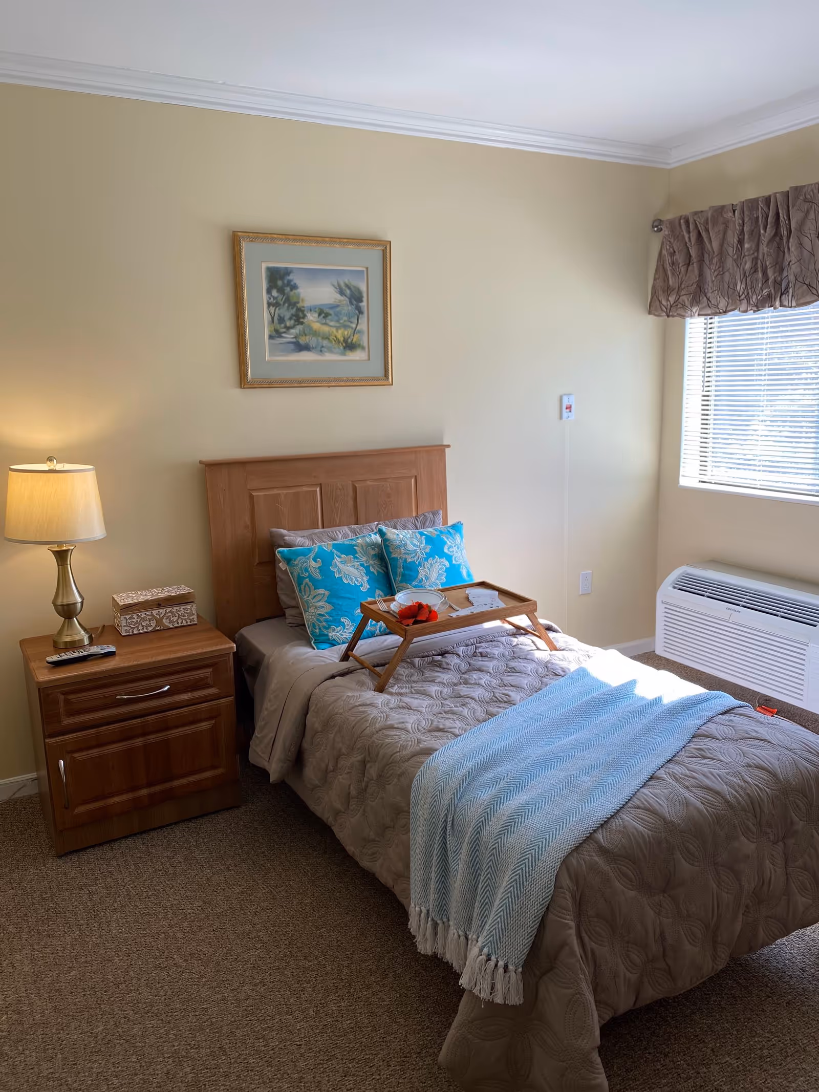A tidy single bedroom with a wooden headboard, nightstand and lamp, decorative pillows and a breakfast tray on the bed by a window.