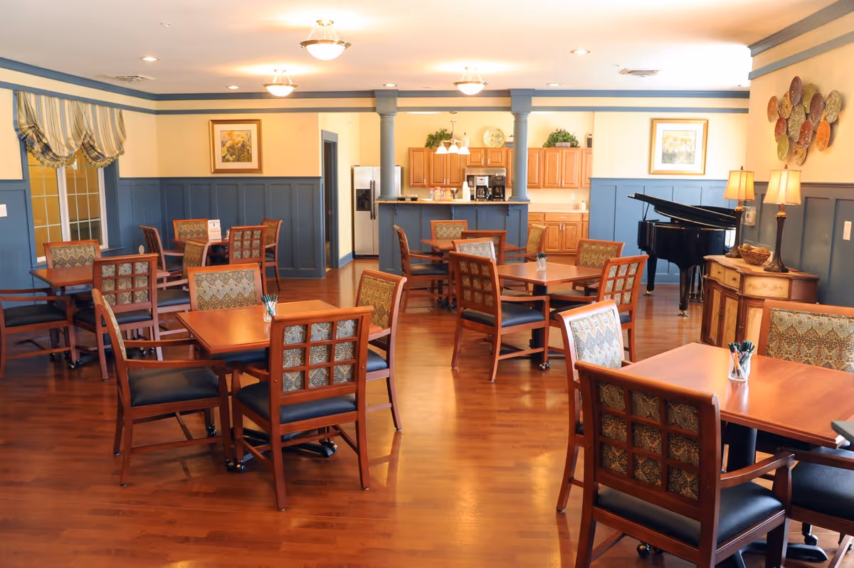 A bright and spacious dining room with multiple wooden tables and chairs arranged neatly on a polished hardwood floor. The walls are painted in a soft yellow with blue wainscoting, decorated with framed floral artwork and a decorative wall piece. In the background, there is a kitchen area with wooden cabinets, a refrigerator, and coffee machines. A black grand piano is positioned near a sideboard with two lamps and a bowl of decorative items.