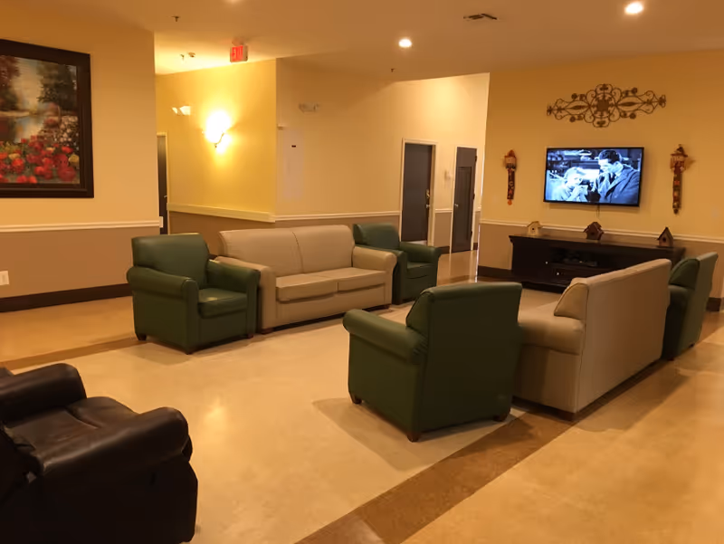 A cozy living room area with beige and green armchairs and sofas arranged around a TV mounted on a wall. The TV is playing a black and white movie. The room has warm yellow walls, a painting of flowers on one wall, and decorative items on a dark wooden TV stand.