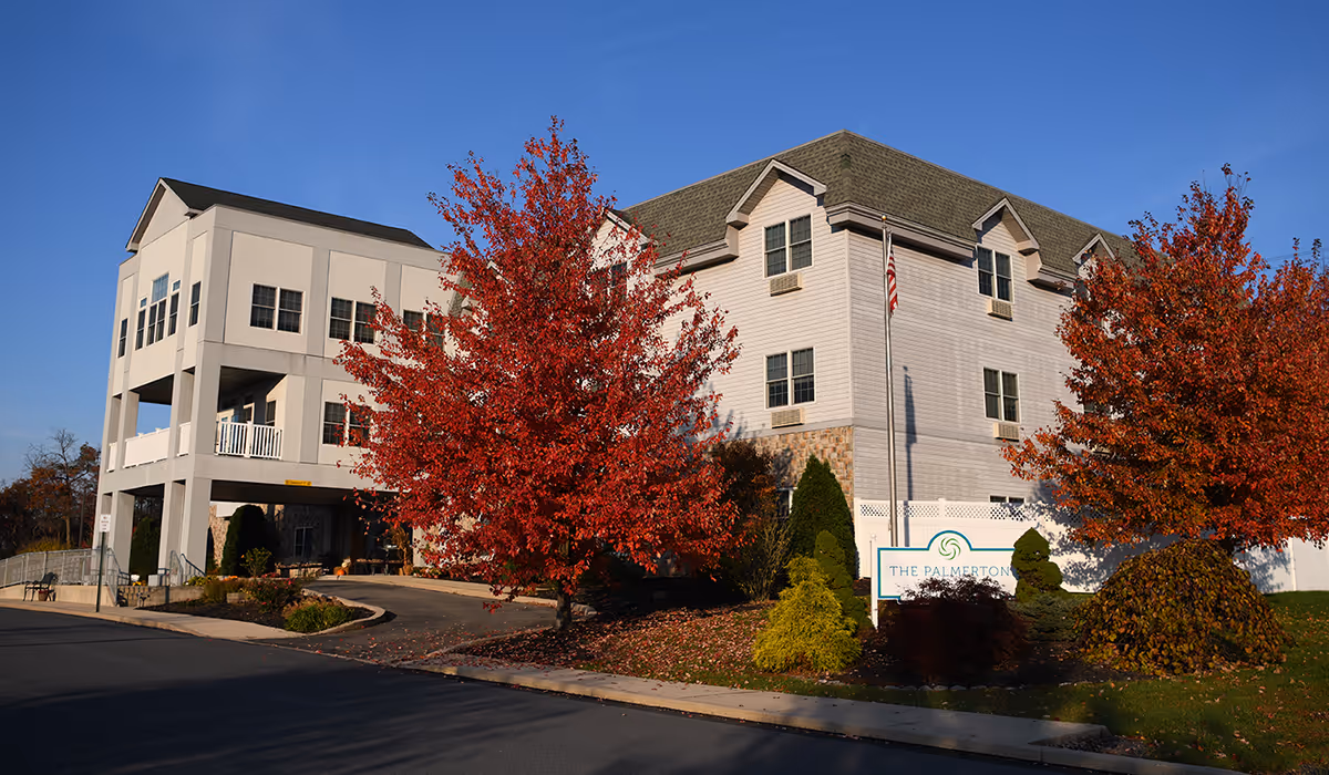 Exterior view of The Palmerton facility building with autumn trees featuring red and orange leaves in front. The building has multiple windows, a covered entrance, and a sign with the facility name near the sidewalk.