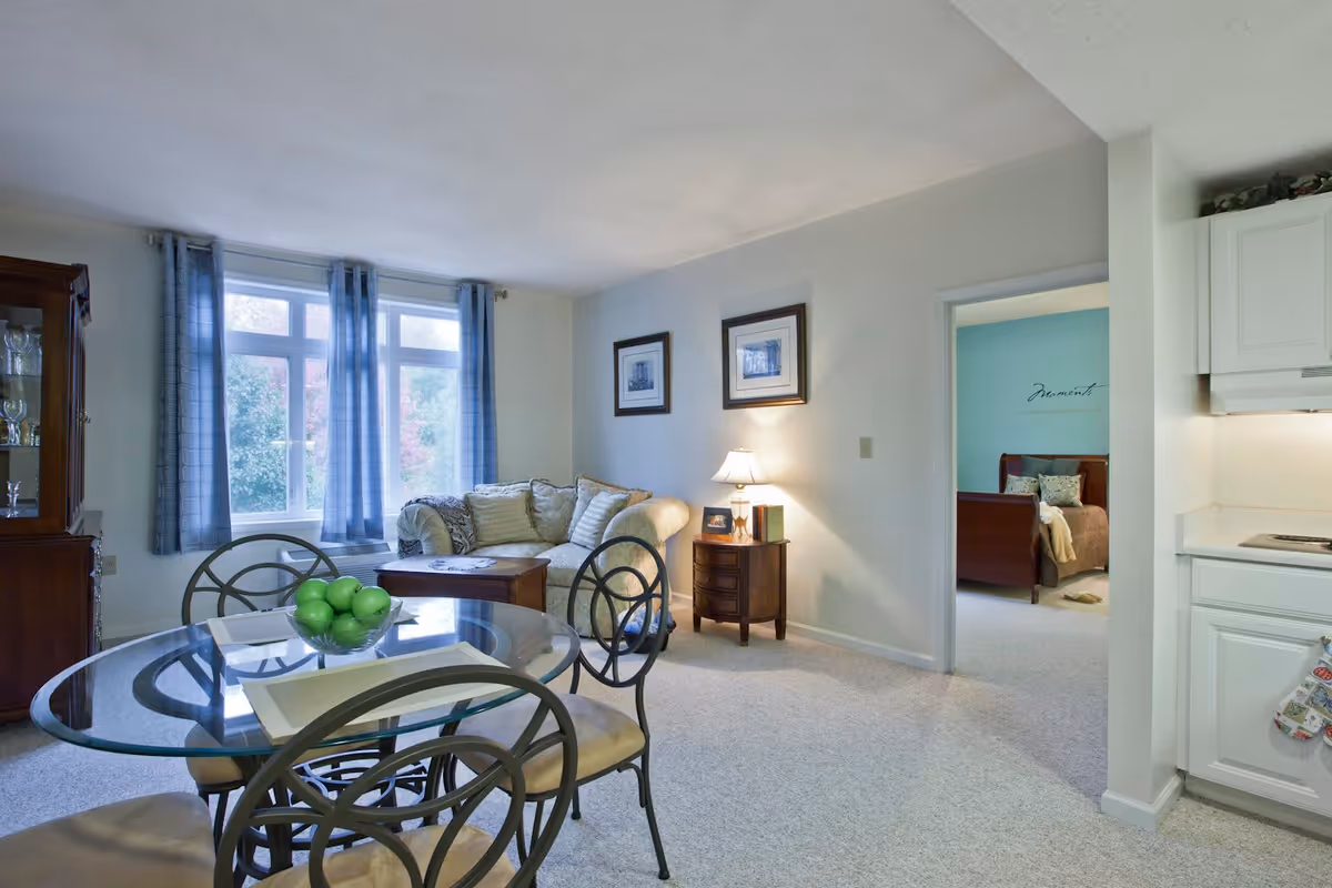 A cozy living and dining area with a glass-top dining table and four chairs in the foreground. Behind the table is a beige couch with patterned cushions, a wooden side table with a lamp, and two framed pictures on the wall. A window with blue curtains lets in natural light. To the right, there is a partial view of a white kitchen and an open doorway leading to a bedroom with a teal accent wall and a wooden bed frame.