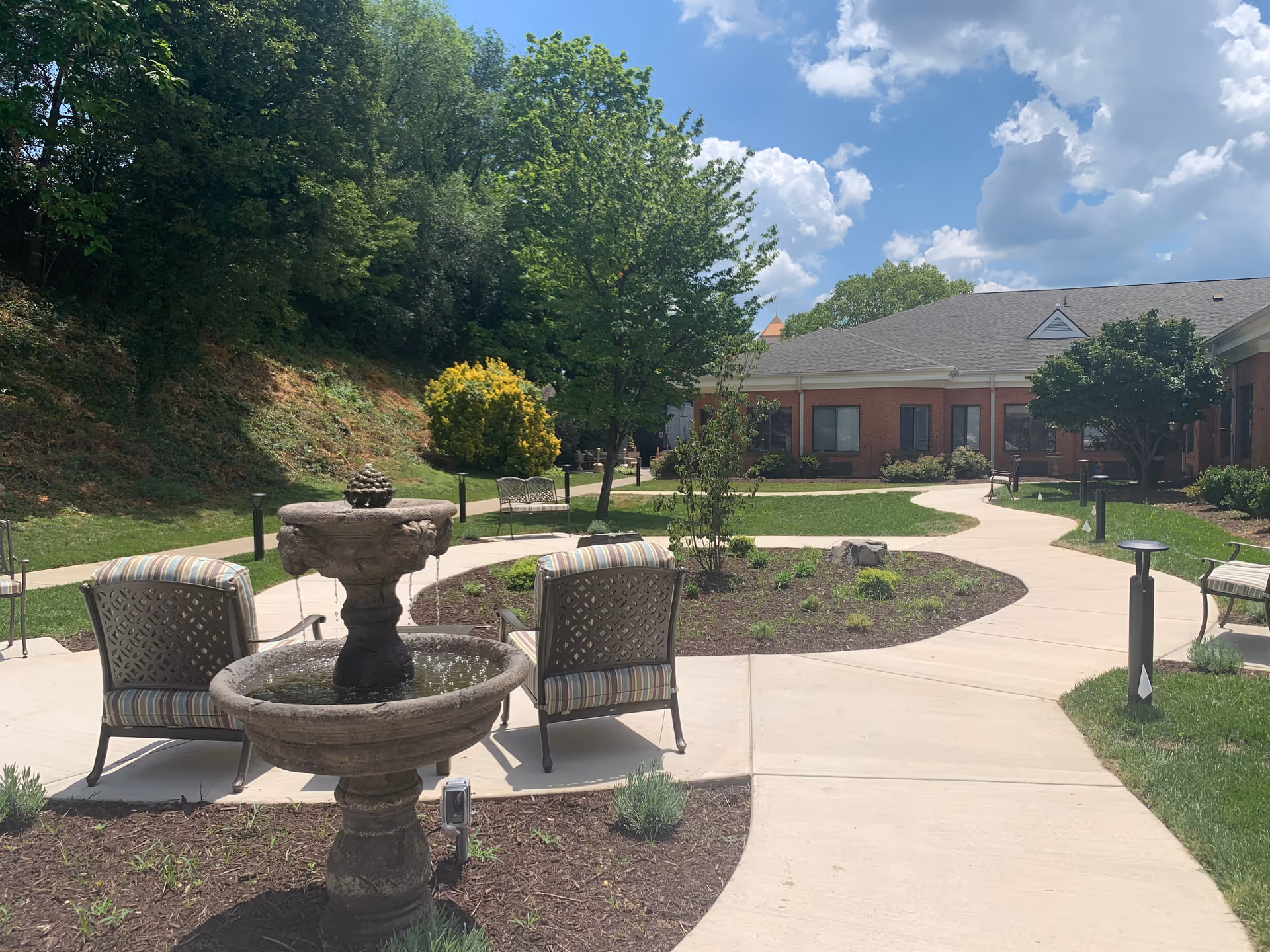 Outdoor courtyard area at a senior living facility with a stone water fountain in the foreground, surrounded by cushioned metal chairs. Curved concrete walkways lead through landscaped garden beds with small plants and trees. A brick building with windows is visible in the background under a partly cloudy blue sky.