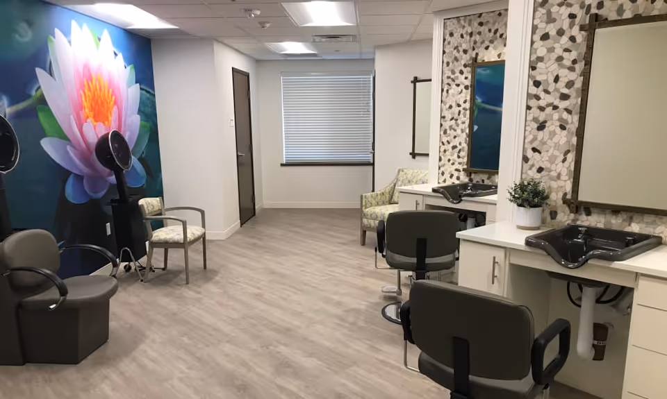 Interior of a salon area in a senior living facility with salon chairs, hair drying stations, sinks, and a large floral mural on one wall. The room has light wood flooring, a window with blinds, and a few additional chairs for seating.