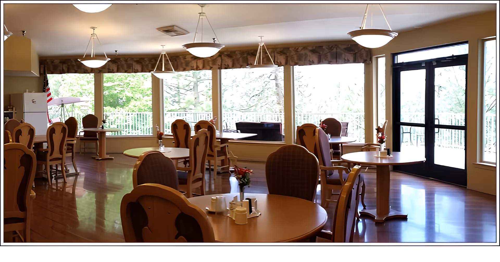 A bright dining room with several round wooden tables and chairs arranged neatly. Large windows along one wall provide a view of green trees outside, and there are hanging ceiling lights illuminating the space. A door with glass panels leads to an outdoor area, and a refrigerator is visible in the corner near an American flag.