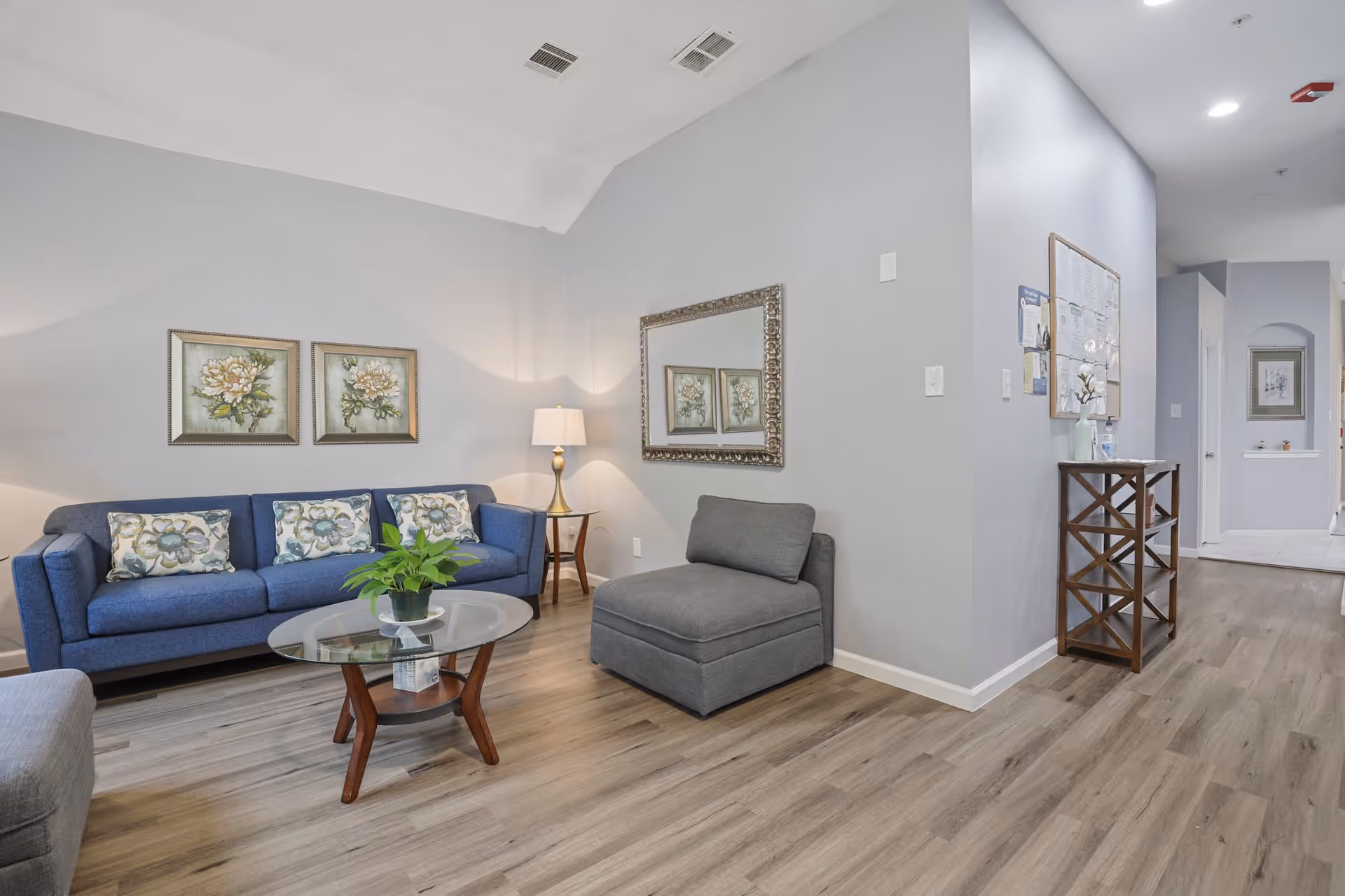 A cozy living room area in a senior living facility featuring a blue sofa with floral cushions, a glass coffee table with a green plant, a gray armchair, a side table with a lamp, two floral paintings on the wall, a large decorative mirror, and a wooden shelving unit against a light gray wall with wood flooring.