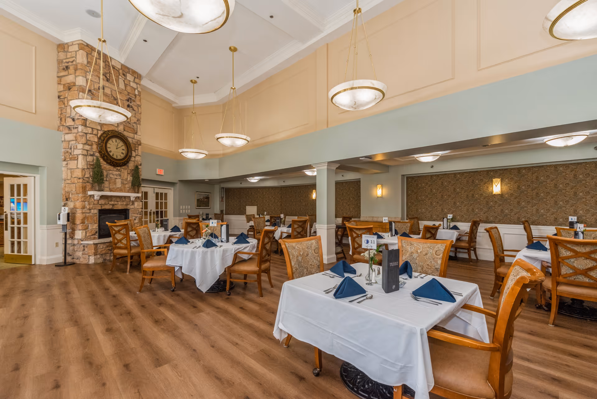 A spacious dining room with wooden floors, multiple tables covered with white tablecloths, each set with blue napkins and silverware. The room features a stone fireplace with a large clock above it, elegant hanging light fixtures, and a mix of wooden chairs with patterned upholstery. The walls are painted in soft beige and light blue tones, with decorative wallpaper on one side.