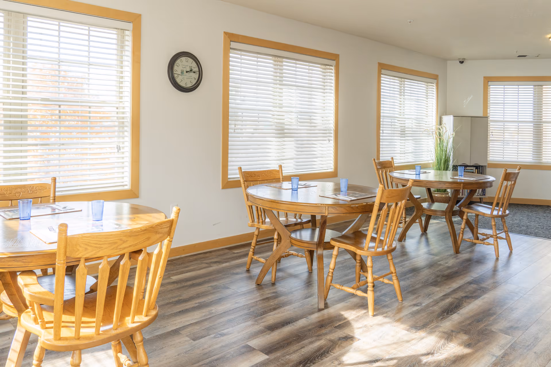 A bright dining room with three round wooden tables, each surrounded by wooden chairs. Each table has placemats and blue drinking glasses. The room has large windows with white blinds, light-colored walls, and wood trim. A clock is mounted on the wall, and there is a plant in the corner near a cabinet.