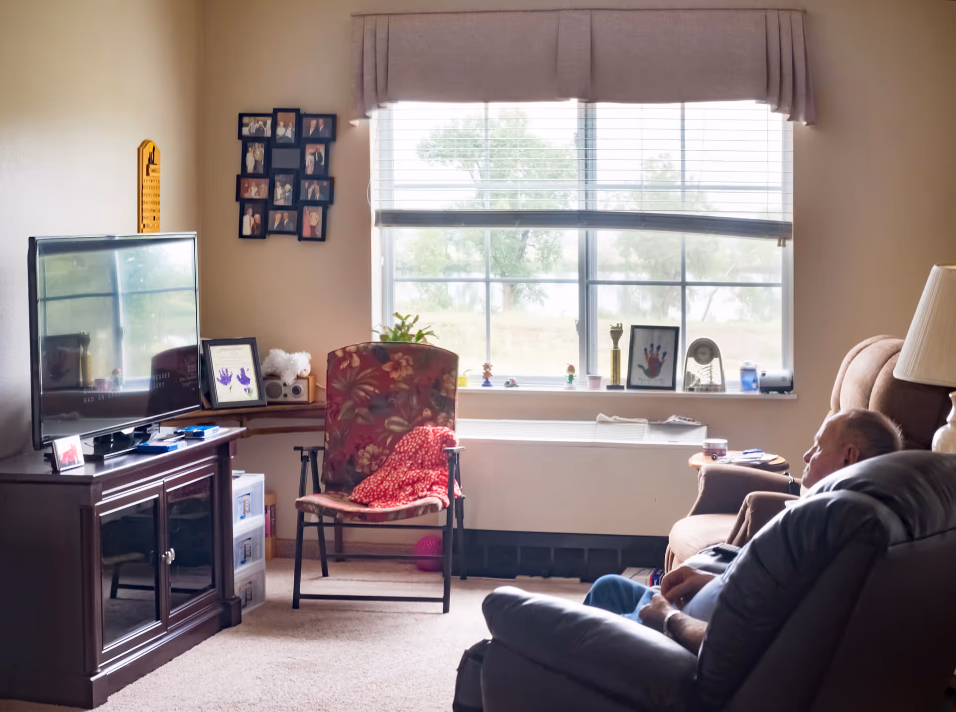A cozy living room in a senior living facility with a large window letting in natural light. There is a floral-patterned chair with a red blanket draped over it, a wooden TV stand with a flat-screen television, and a man sitting in a recliner chair watching TV. The window sill has various small decorative items and framed pictures. A lamp is positioned next to the recliner.