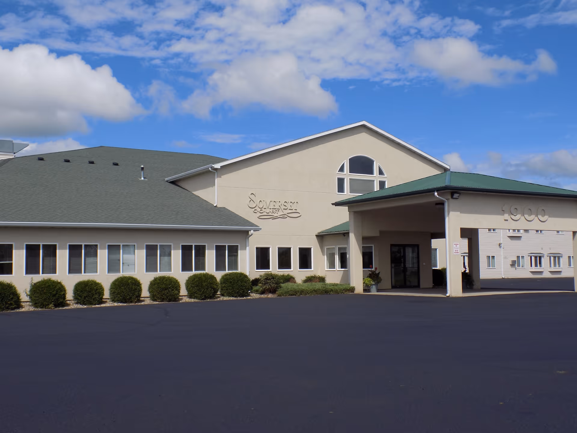 Exterior view of Somerset Court senior living facility building with a green roof, beige walls, multiple windows, and an entrance canopy. The sky is partly cloudy and the parking area is visible in front.