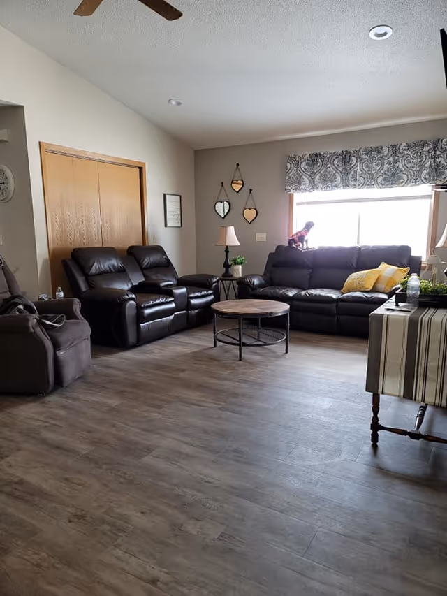 A cozy living room with dark leather sofas and recliners arranged around a round wooden coffee table. There is a window with patterned valance letting in natural light, and a small dog sitting on one of the sofas. The walls are decorated with three heart-shaped mirrors and a framed picture. A side table with a lamp and a small plant is next to the sofa, and another table with a striped tablecloth is partially visible on the right.
