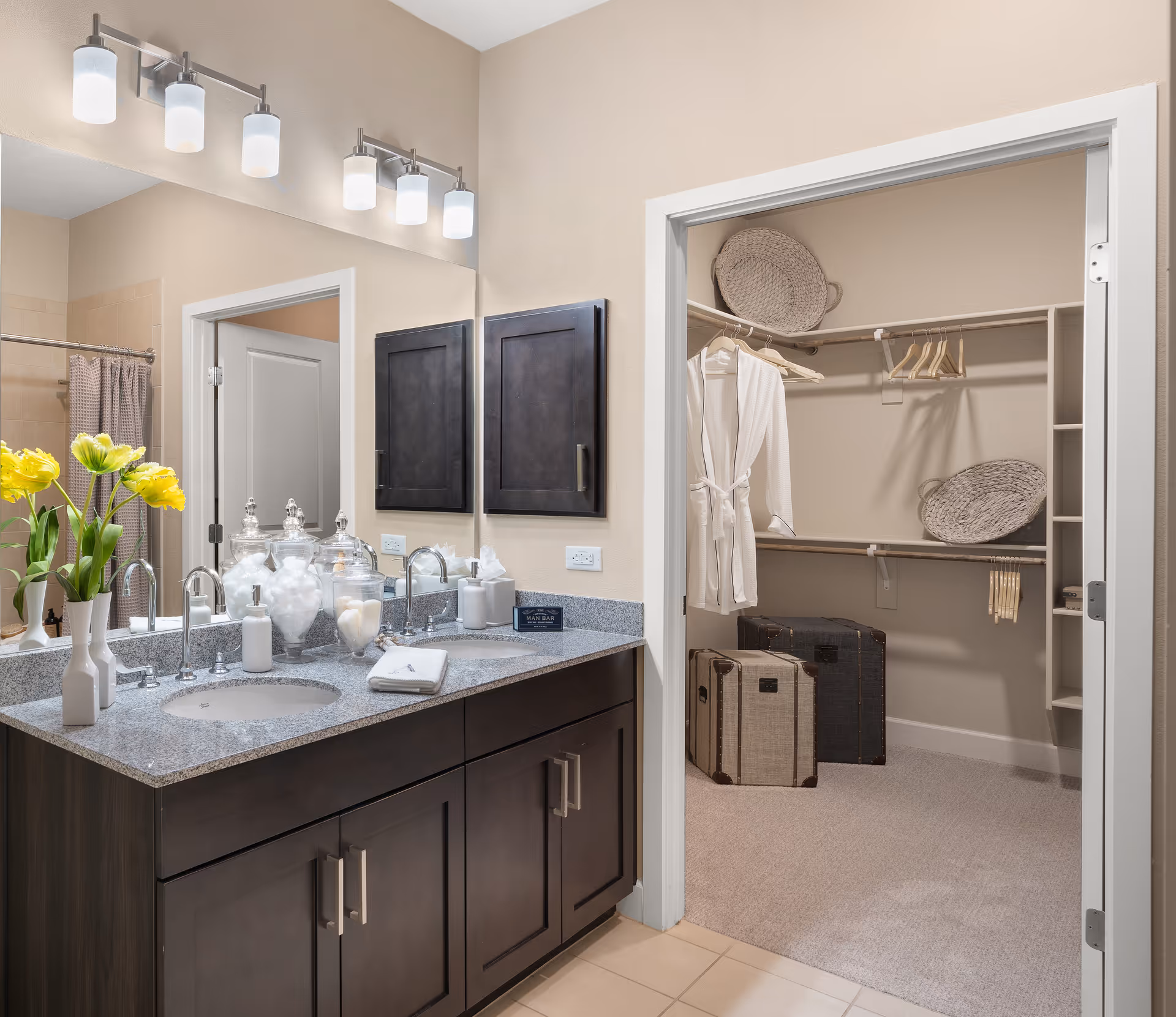 A bathroom with a double sink vanity featuring dark wood cabinets and a granite countertop. Above the sinks are two sets of three-light fixtures and a large mirror. On the countertop are decorative jars, a soap dispenser, a tissue box, and a small towel. To the right is an open doorway leading to a walk-in closet with hanging robes, wicker baskets, and two suitcases on the carpeted floor.