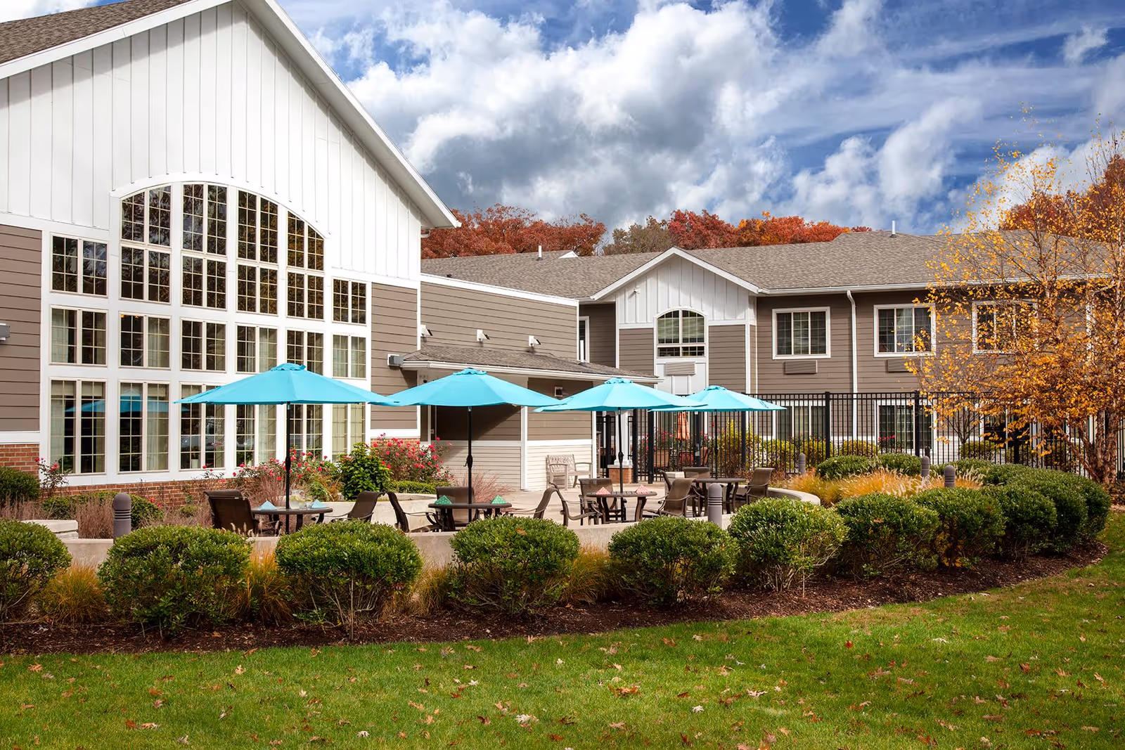 Outdoor patio area of a senior living facility with several tables and chairs under blue umbrellas, surrounded by neatly trimmed bushes and landscaping. The building has large windows and a mix of white and beige siding with a sloped roof. Trees with autumn foliage are visible in the background under a partly cloudy sky.