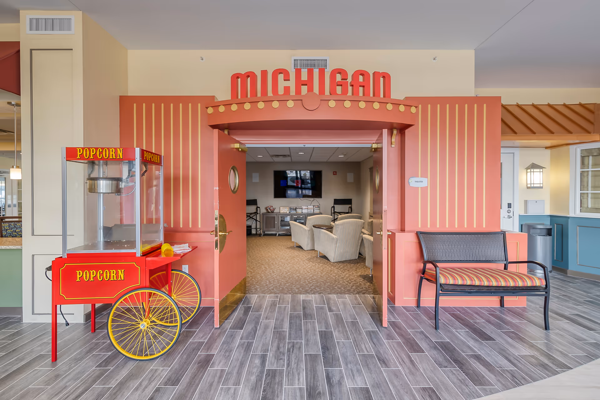 Entrance to a theater room labeled 'Michigan' with double red doors open to a seating area with armchairs and a TV. To the left of the entrance is a red and yellow popcorn cart, and to the right is a bench with a striped cushion. The floor is tiled in gray wood-like planks.