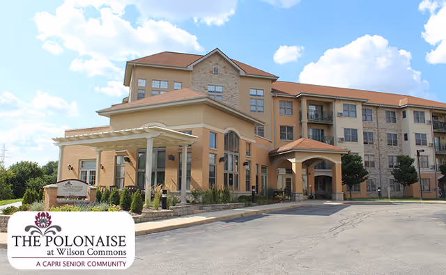 Exterior view of a senior living facility building with multiple floors, beige and stone facade, and a covered entrance. The sky is partly cloudy and there is a paved driveway in front.