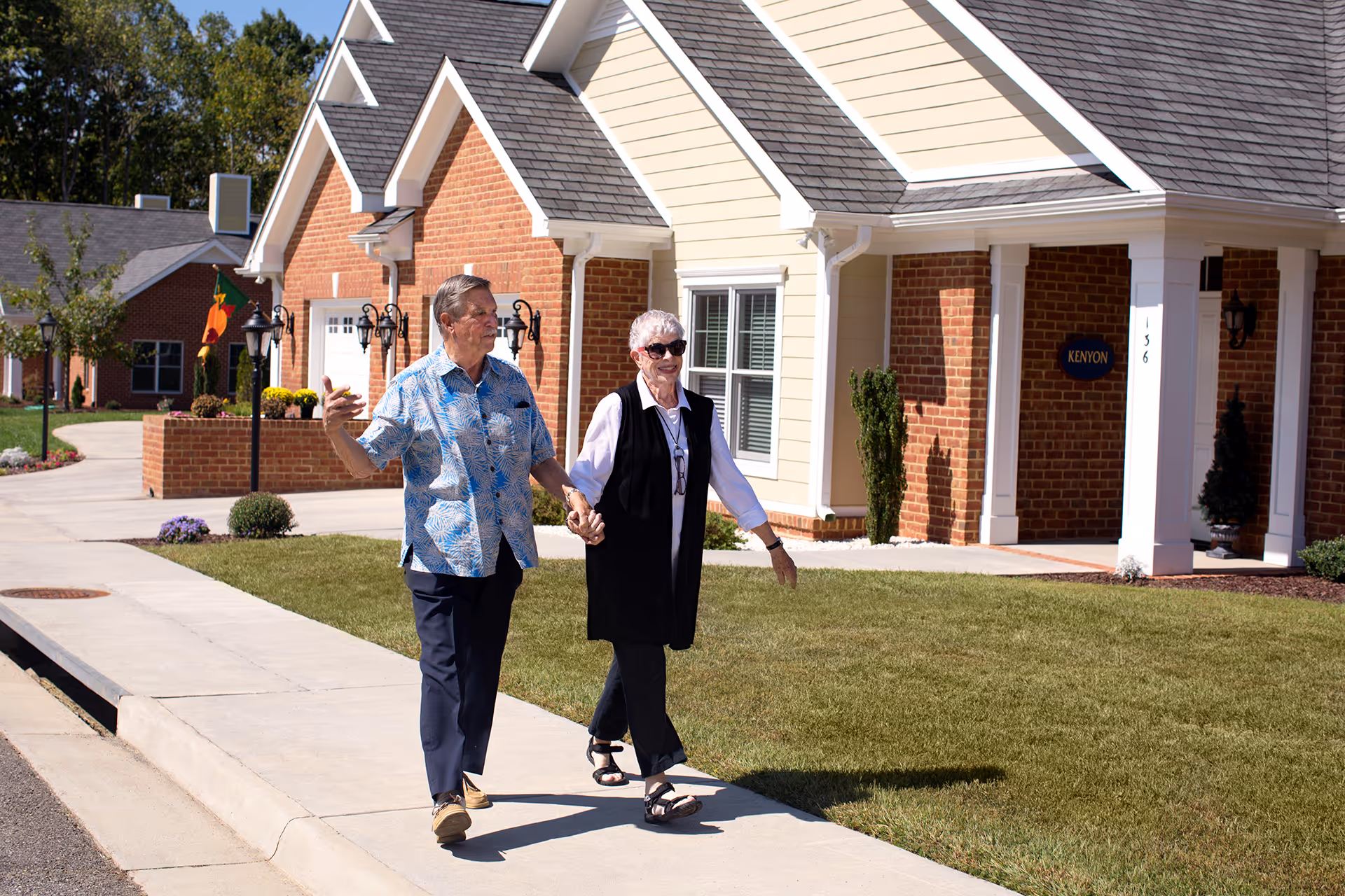 An elderly couple holding hands and walking on a sidewalk in front of a residential building with brick and light-colored siding. The man is wearing a blue patterned shirt and dark pants, and the woman is wearing a black vest over a white shirt with black pants and sunglasses. The building has a sign that reads 'KENYON' and the number 136 on a column.