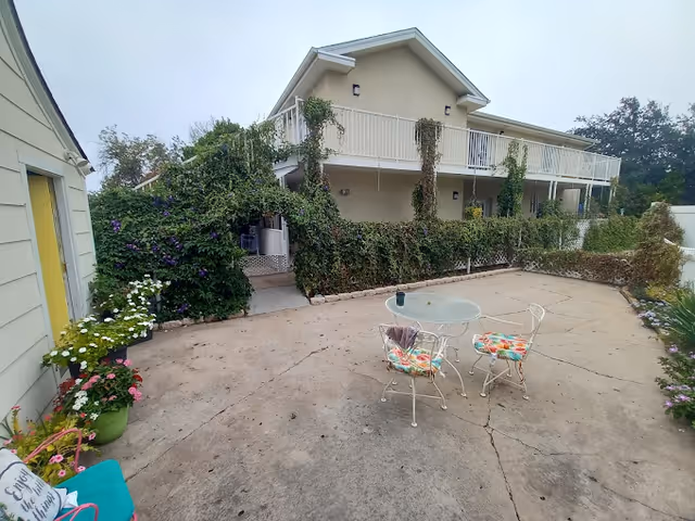 Outdoor patio area with a round glass table and two metal chairs with colorful cushions. The patio is surrounded by greenery, including flowering plants and vines climbing a two-story building with a balcony. The sky is overcast.