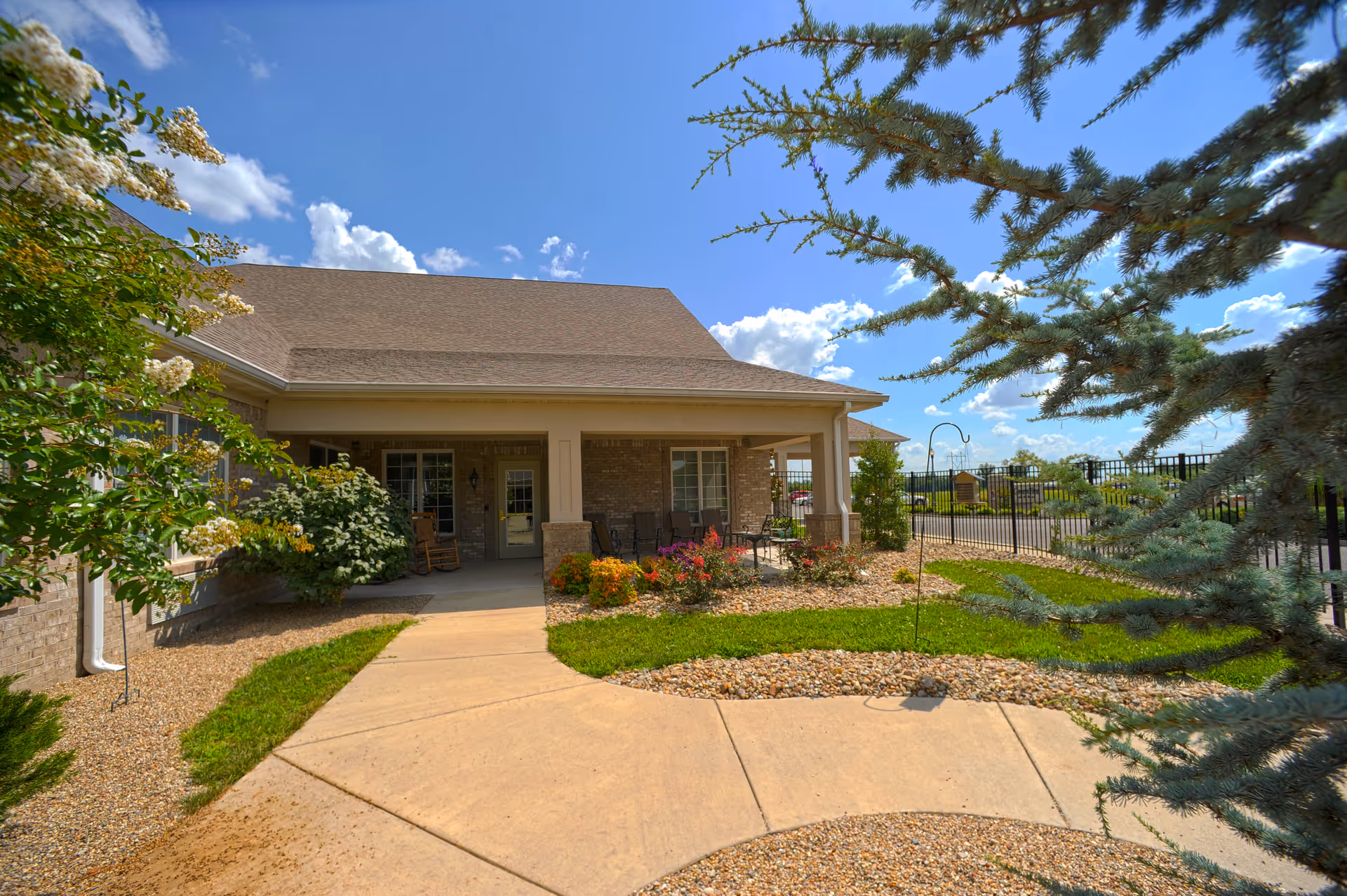 Exterior view of a single-story brick building with a covered porch featuring chairs and a small table. The surrounding area includes a paved walkway, green grass, flowering plants, and trees under a blue sky with scattered clouds.