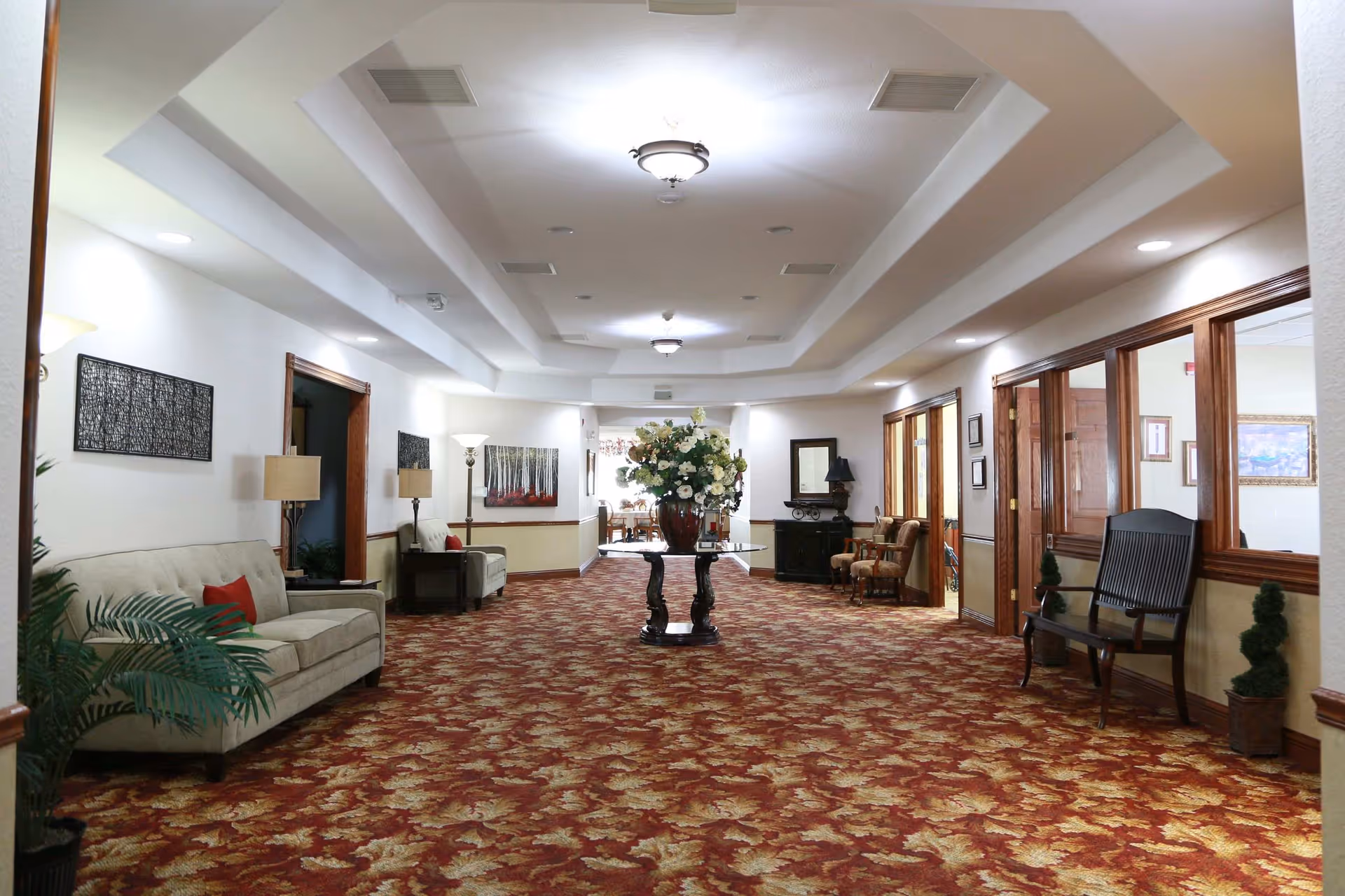 Long carpeted interior hallway with sofas, chairs, a central table of flowers, and framed artwork.