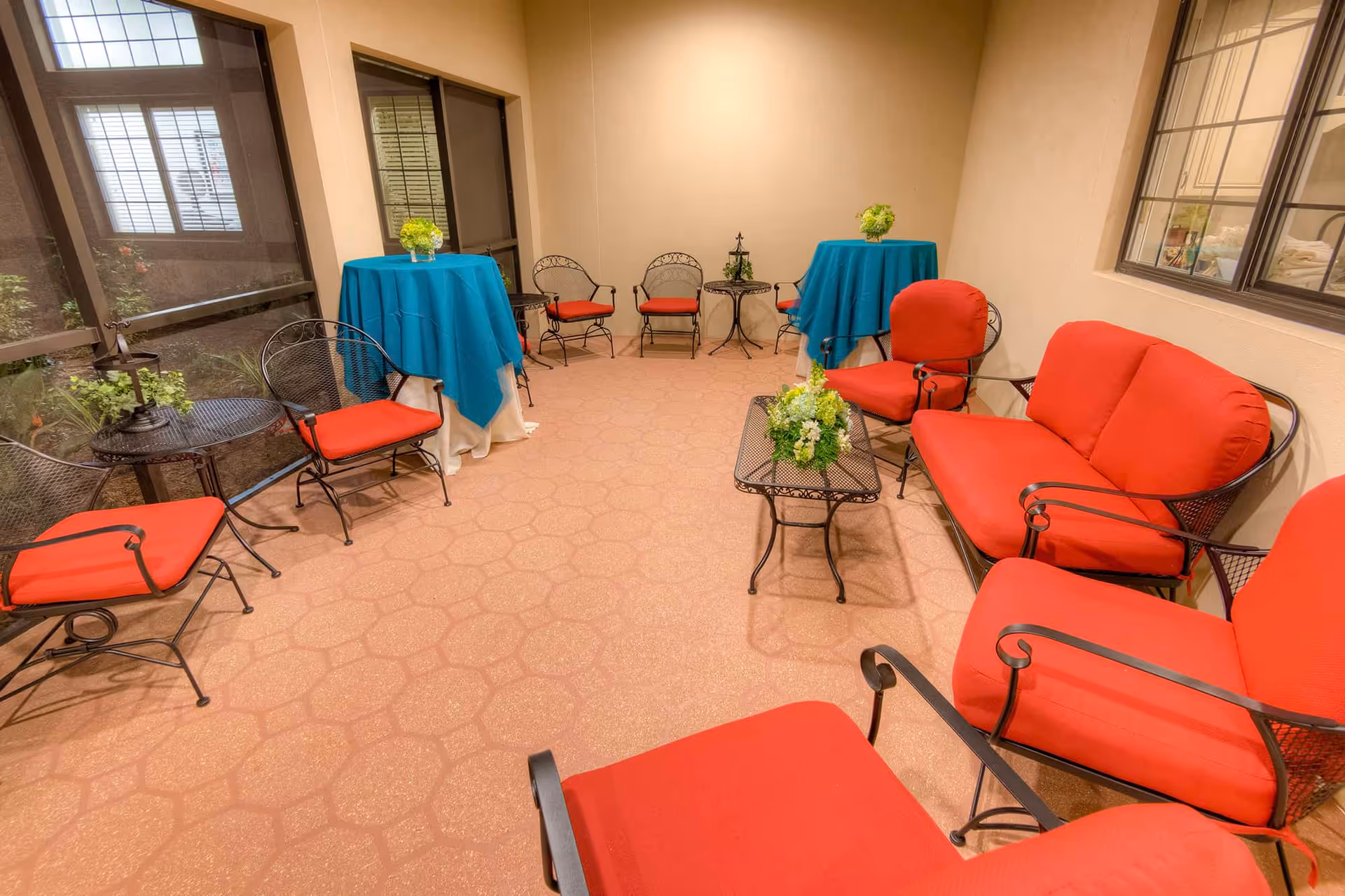 Enclosed indoor seating area with red-cushioned metal chairs and loveseats arranged around small metal tables and turquoise-draped tall tables.