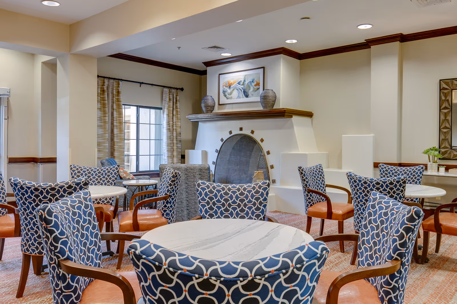 A cozy common area with multiple round tables surrounded by blue patterned chairs with wooden armrests. The room features a decorative fireplace with two vases and a framed abstract painting above it. There is a window with curtains on the left side and a mirror with a geometric frame on the right wall.